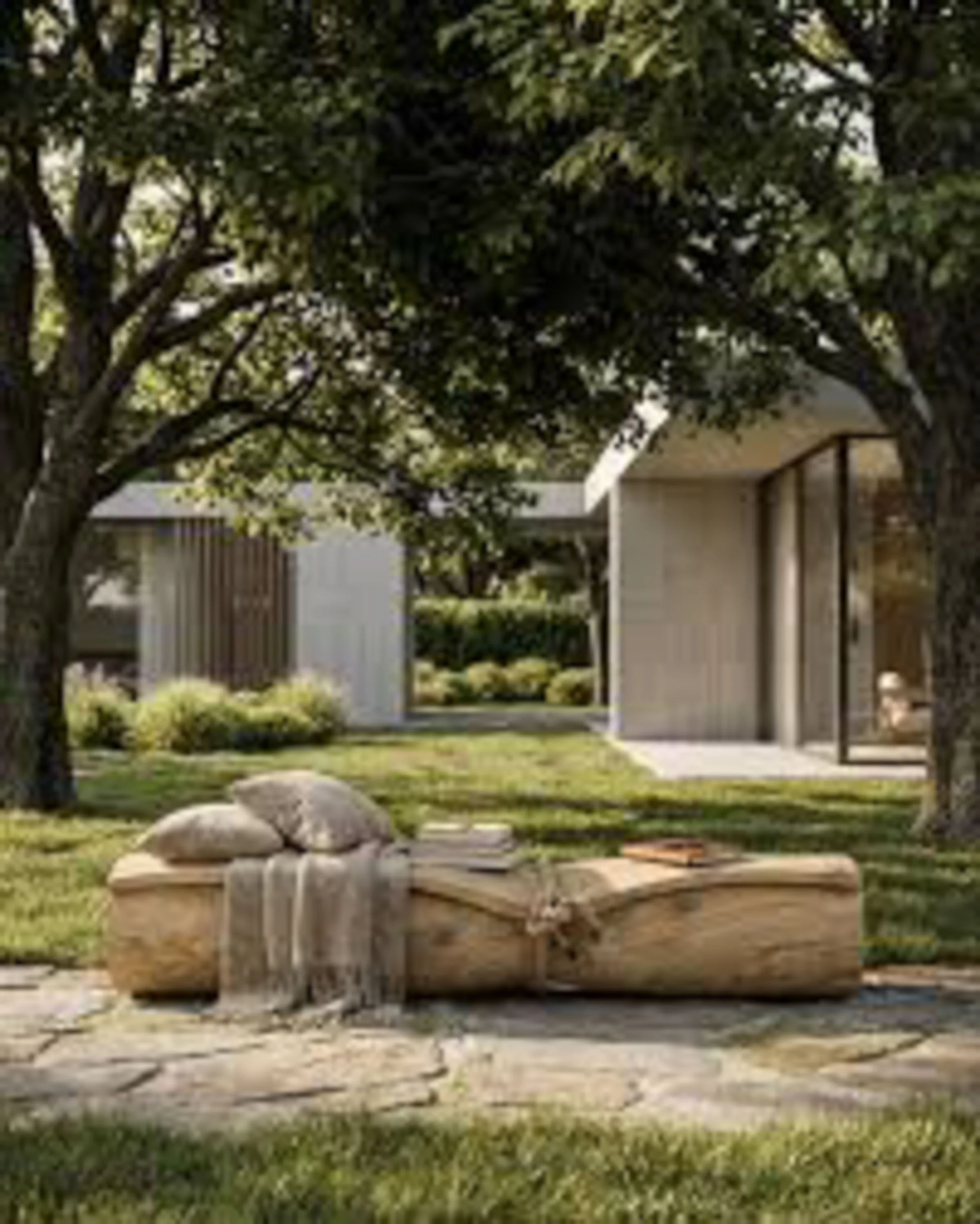 A residential backyard with two trees, a grassy lawn, a stone bench, and a backdrop of a house with a patio covering.