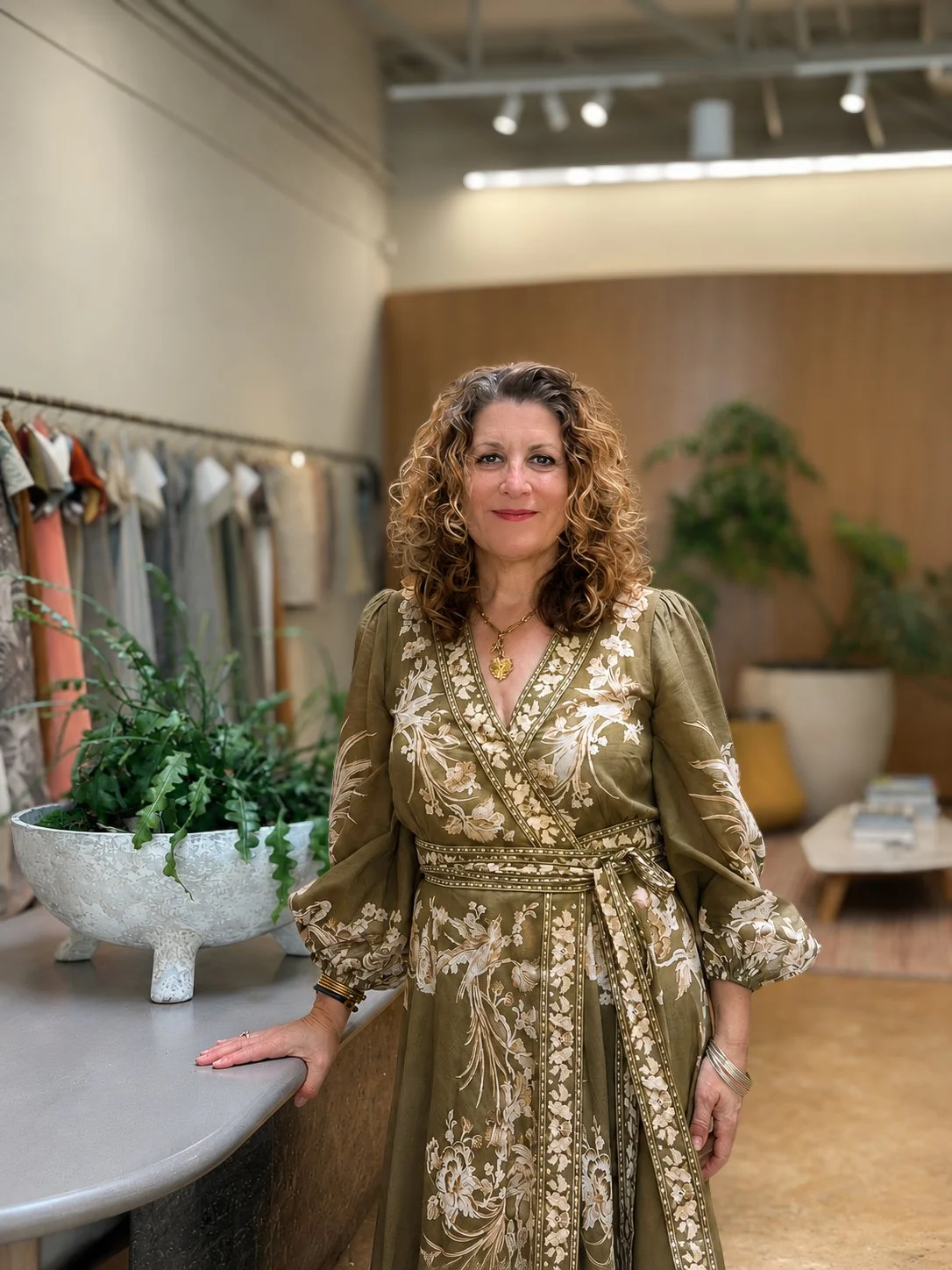 A woman with curly brown hair wearing an ornate olive green dress with floral embroidery, standing indoors in a showroom or boutique with clothing racks and plants in the background.