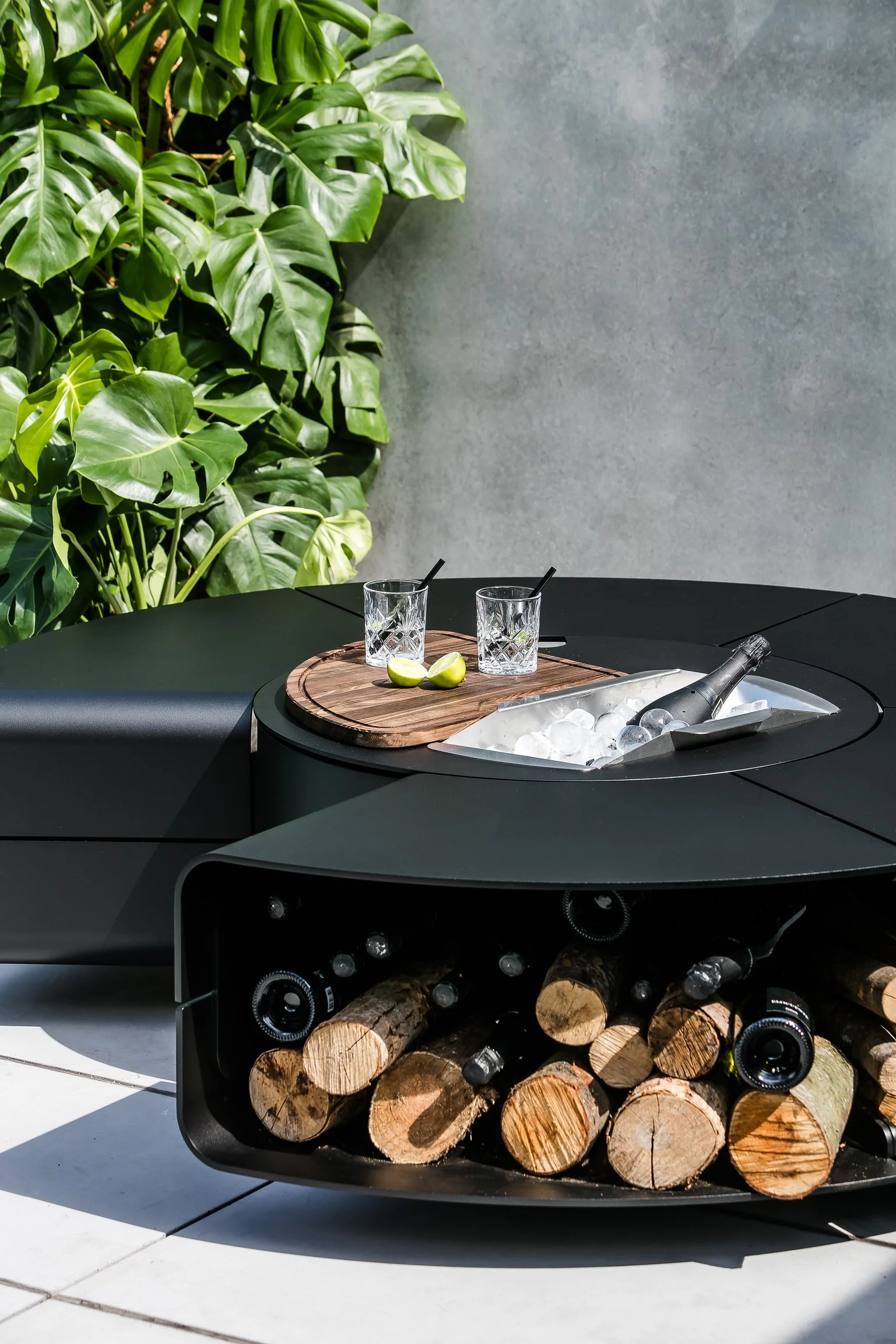 A round black outdoor fire pit with stacked logs inside, topped with a metal grate and two glasses with black straws on a wooden serving tray, set against a gray wall with green leafy plants to the left.