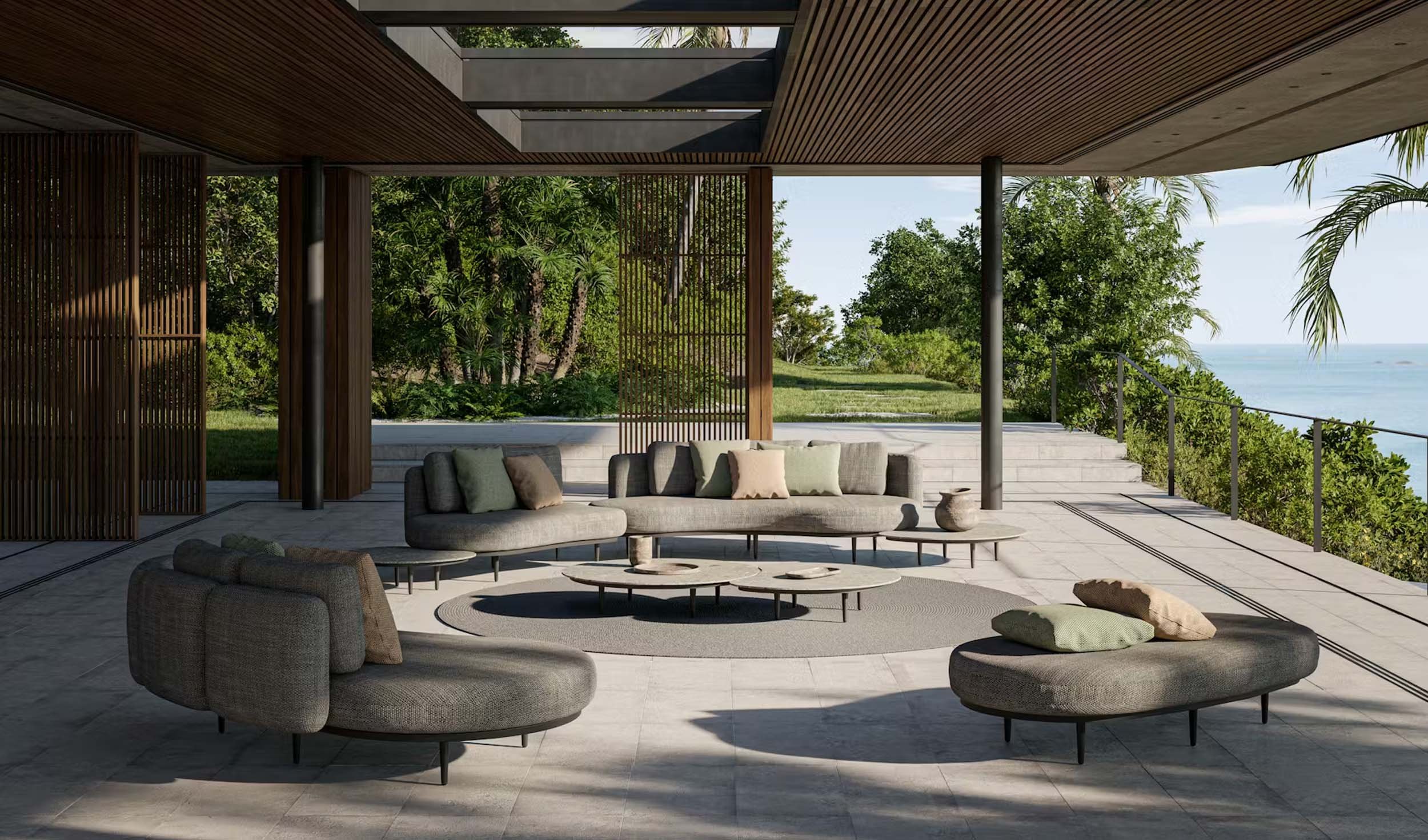 Open-air modern living space with gray sofas and pillows, round coffee tables, and a view of lush greenery and the ocean in the background.