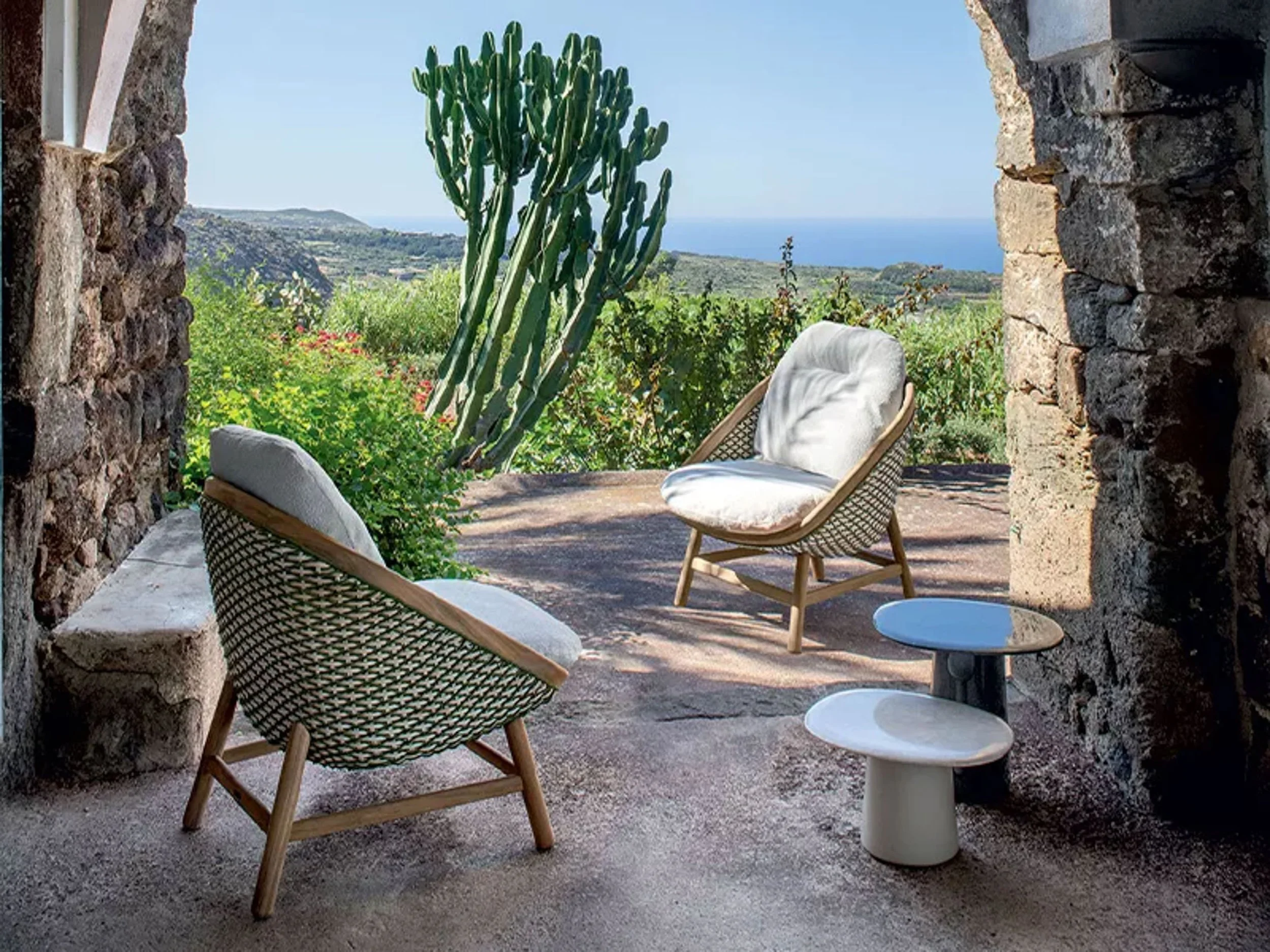 Outdoor seating area with wicker chairs, small white side tables, and a large cactus, overlooking a scenic landscape with hills and ocean in the distance.