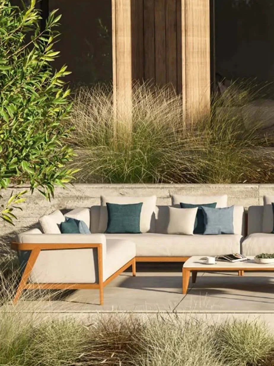 Outdoor patio with a white sofa and blue cushions, coffee table with magazines, surrounded by plants and grass, with a wood fence in the background.