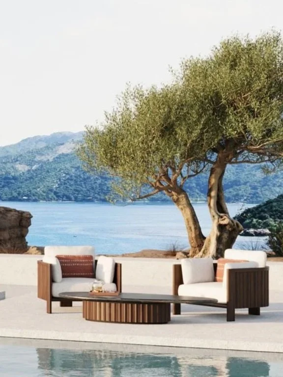 Outdoor seating area with modern white sofas, a wooden coffee table, and a large tree overlooking a body of water with mountains in the background.