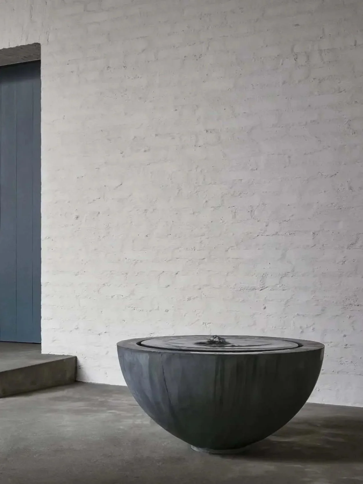 A large, round, black marble fountain on a concrete floor against a white brick wall. To the left, part of a blue door and concrete steps are visible.
