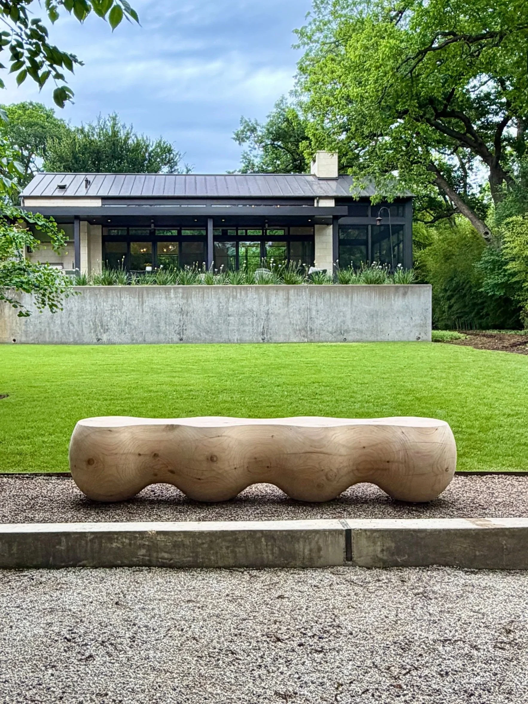 A wooden bench with a rounded, organic shape is placed on a gravel surface in an outdoor garden setting, with lush green trees and grass in the background.