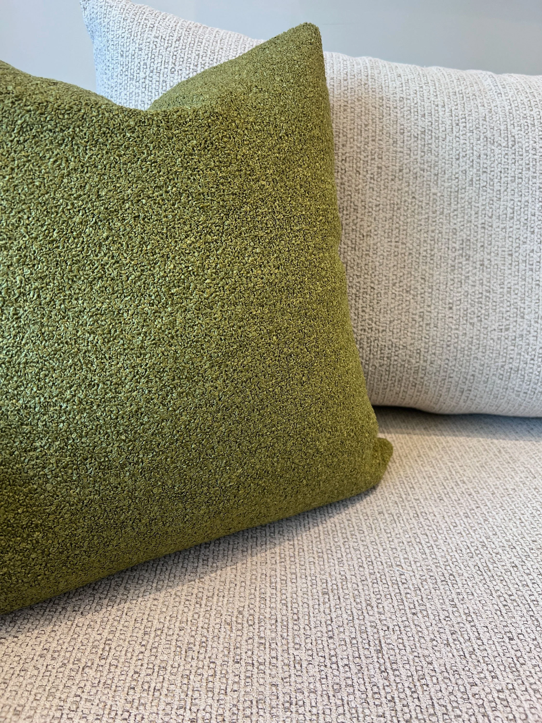 Close-up of two decorative pillows, one olive green with a textured fabric, the other off-white with a subtle pattern, placed on a light-colored textured fabric sofa.