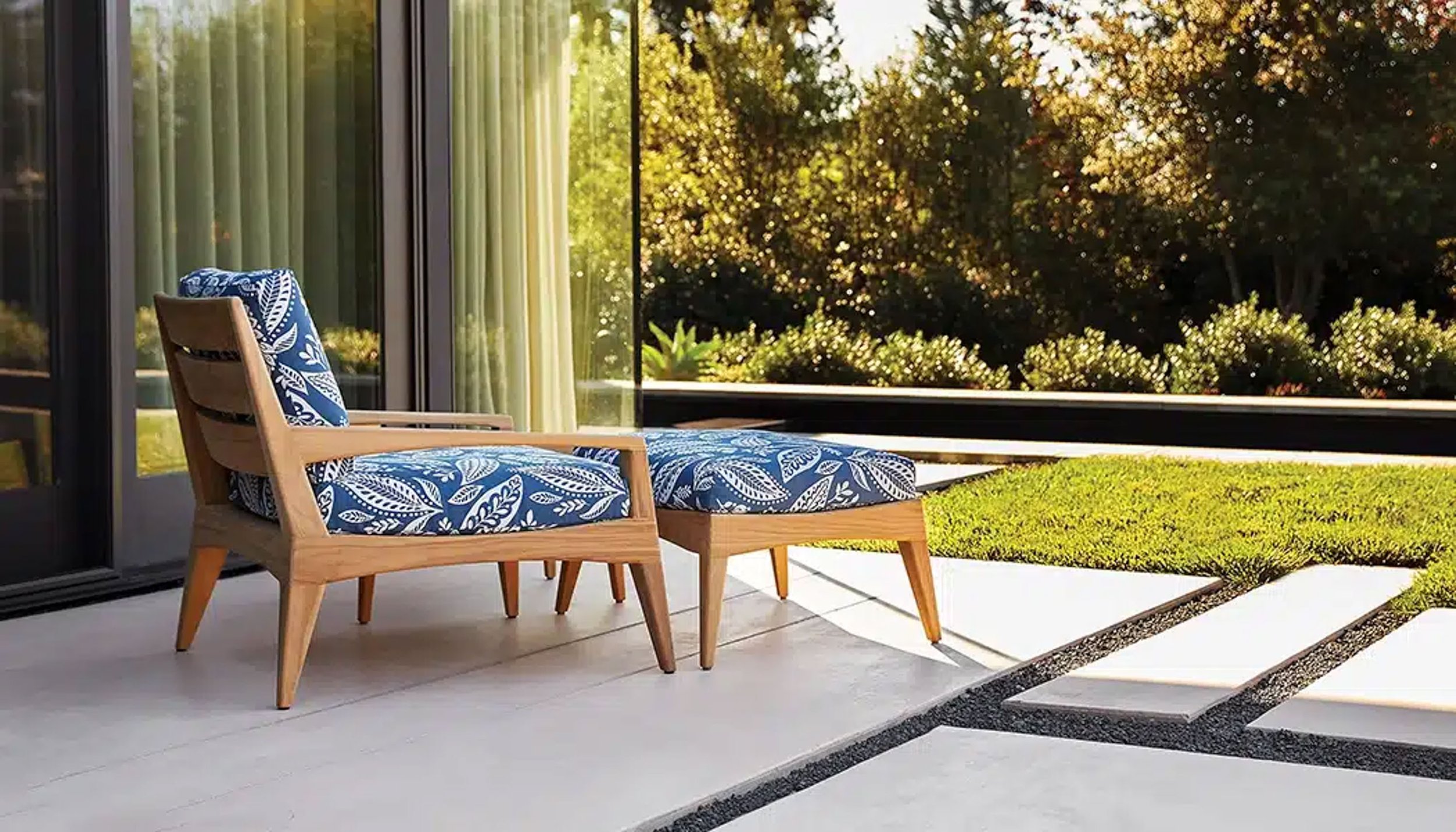 A wooden lounge chair with a blue and white patterned cushion and pillow on a patio. There are sliding glass doors in the background with sheer curtains, and a lush green garden with trees and bushes beyond.
