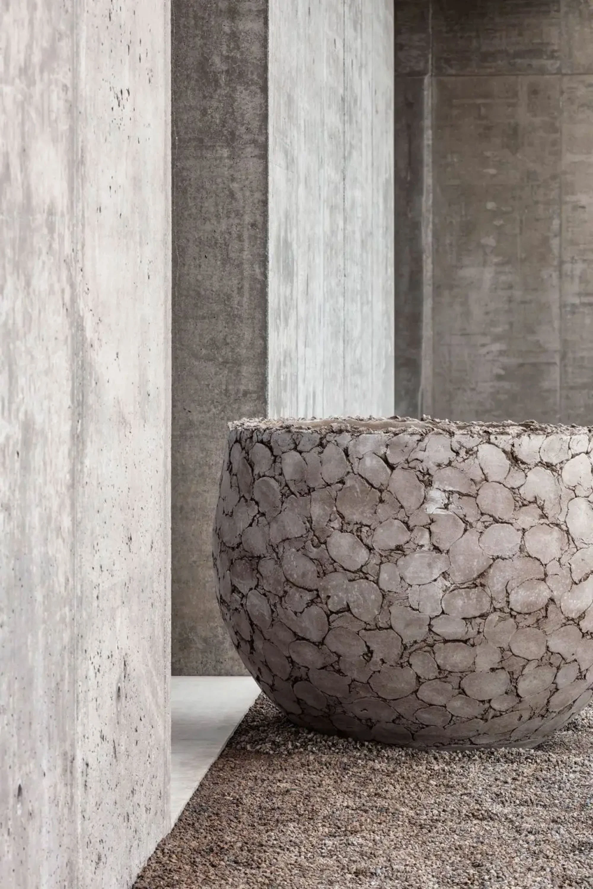 Close-up of a large, round, textured stone planter filled with soil, positioned on gravel ground next to a beige concrete wall with multiple textured sections.