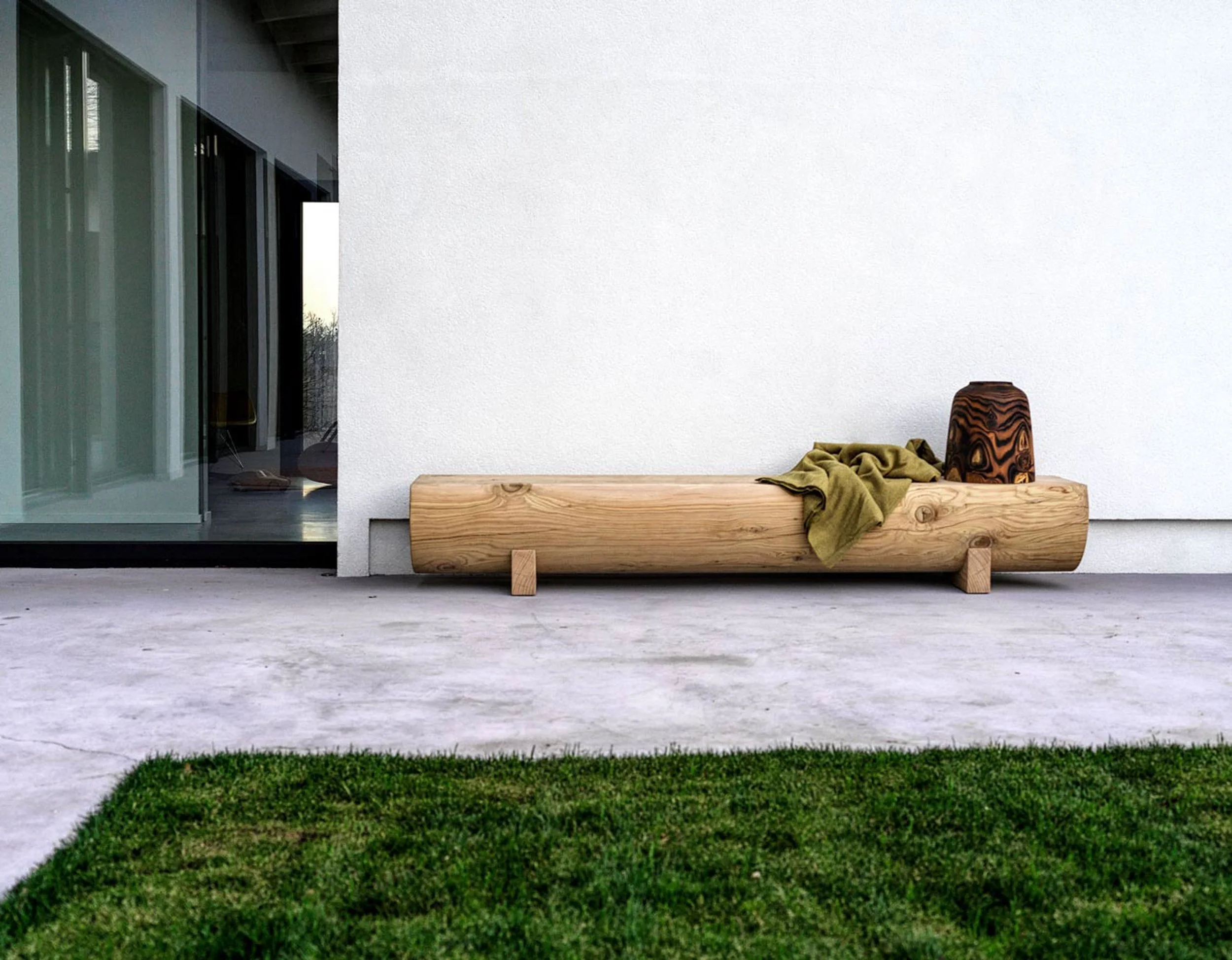 A modern outdoor porch with a wooden bench and a small decorated wooden stool against a white textured wall, with a sliding glass door on the left and a small patch of grass in the foreground.