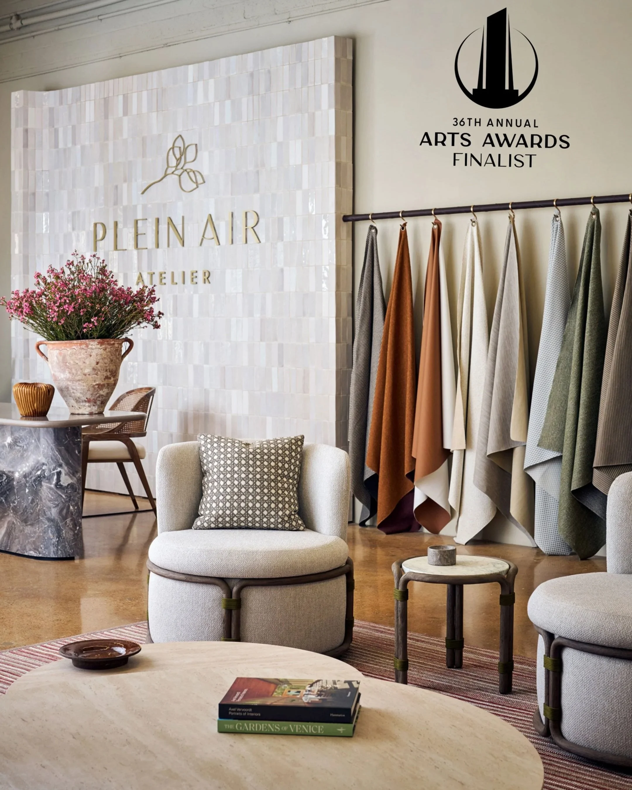 Interior of a modern stylish living space with a white armchair, small side table, and coffee table. The background features fabric swatches hanging on a rack, a wall with logo and text relating to arts awards, and a large potted fern on a marble table. The room has a warm tone with wooden flooring and decorative accents.