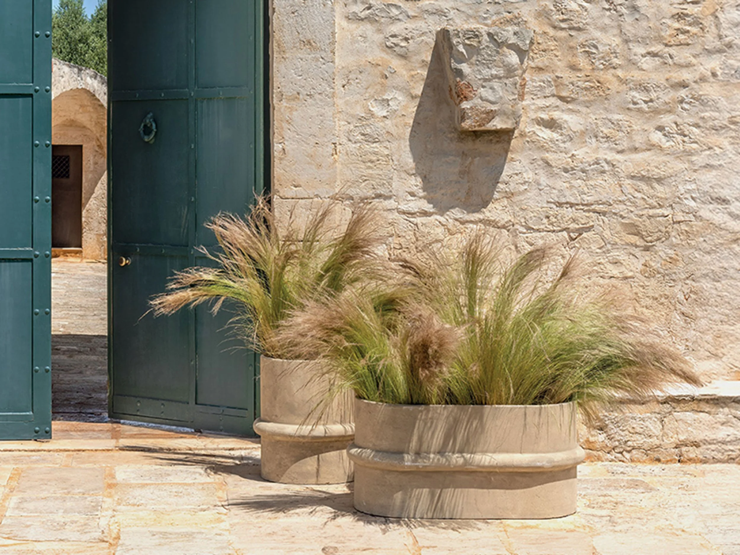 Large planter with ornamental grass in front of stone wall and open green shutters