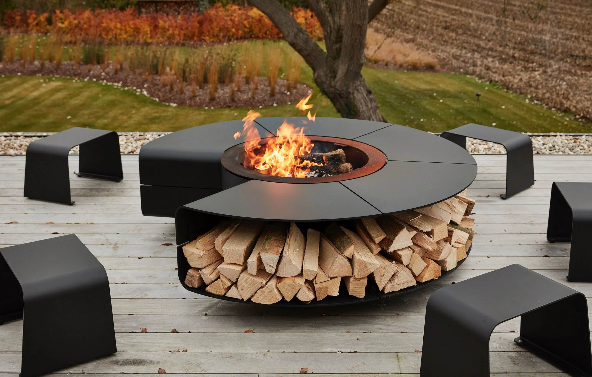 An outdoor fire pit with burning logs, surrounded by black seating, on a wooden deck with a backyard featuring trees and what appears to be a garden in the background.