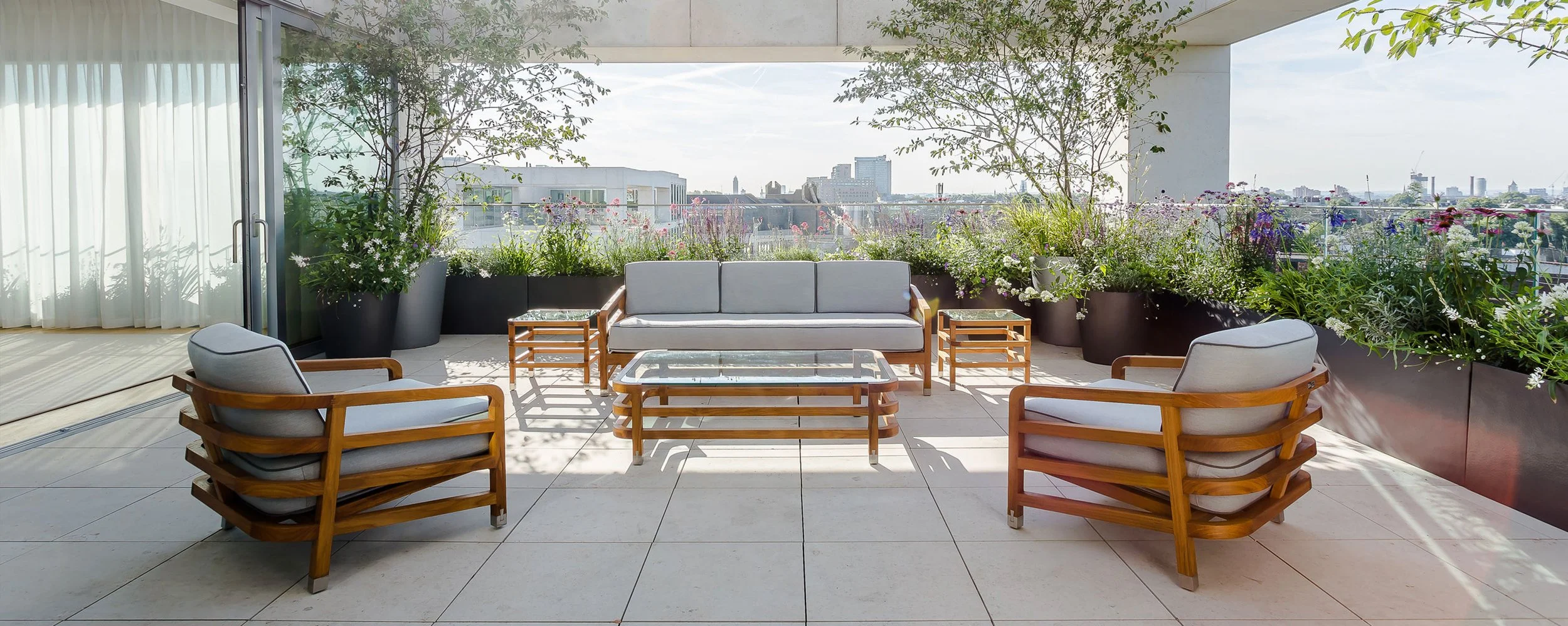 Outdoor terrace with wooden furniture, a cushioned sofa, armchairs, and a central glass coffee table, surrounded by potted flowers and balcony railing overlooking city skyline