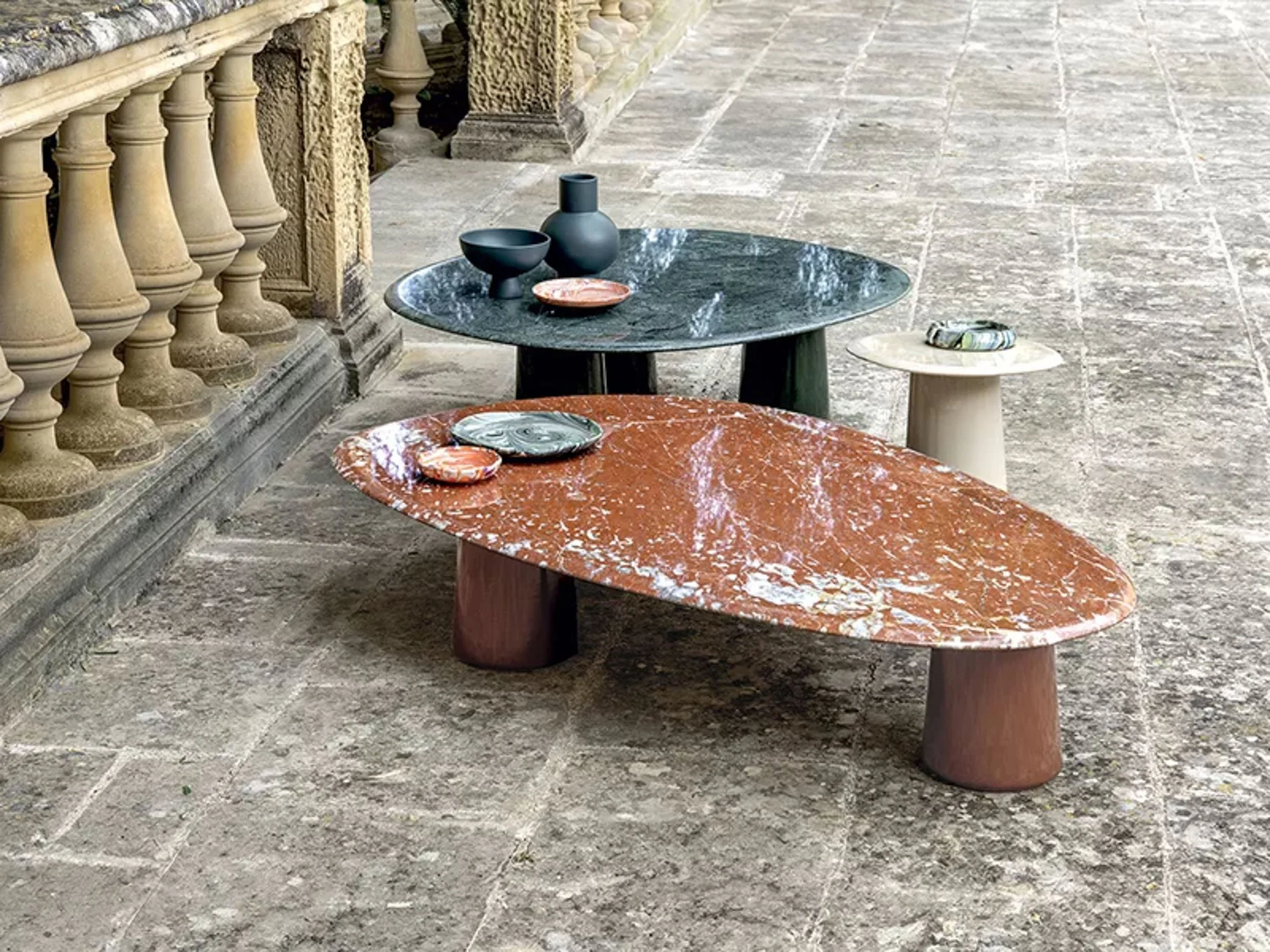 Outdoor stone terrace with a round red marble table and a rectangular green marble table, both decorated with decorative bowls and plates. Stone balustrade and ornate stone wall in the background.