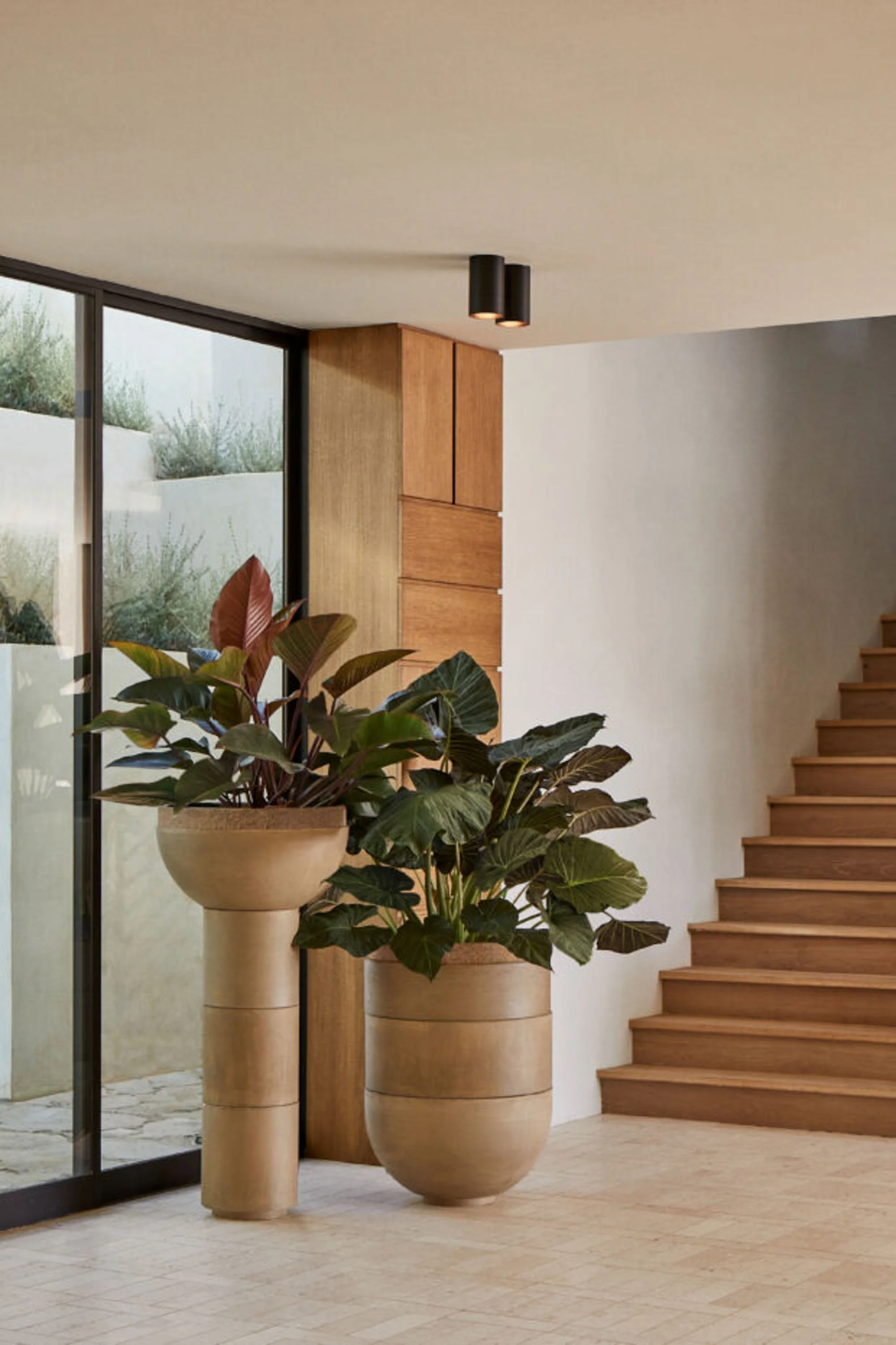 Interior view of a modern home with large potted plants, a wooden staircase, and a floor-to-ceiling window showing an outdoor garden.