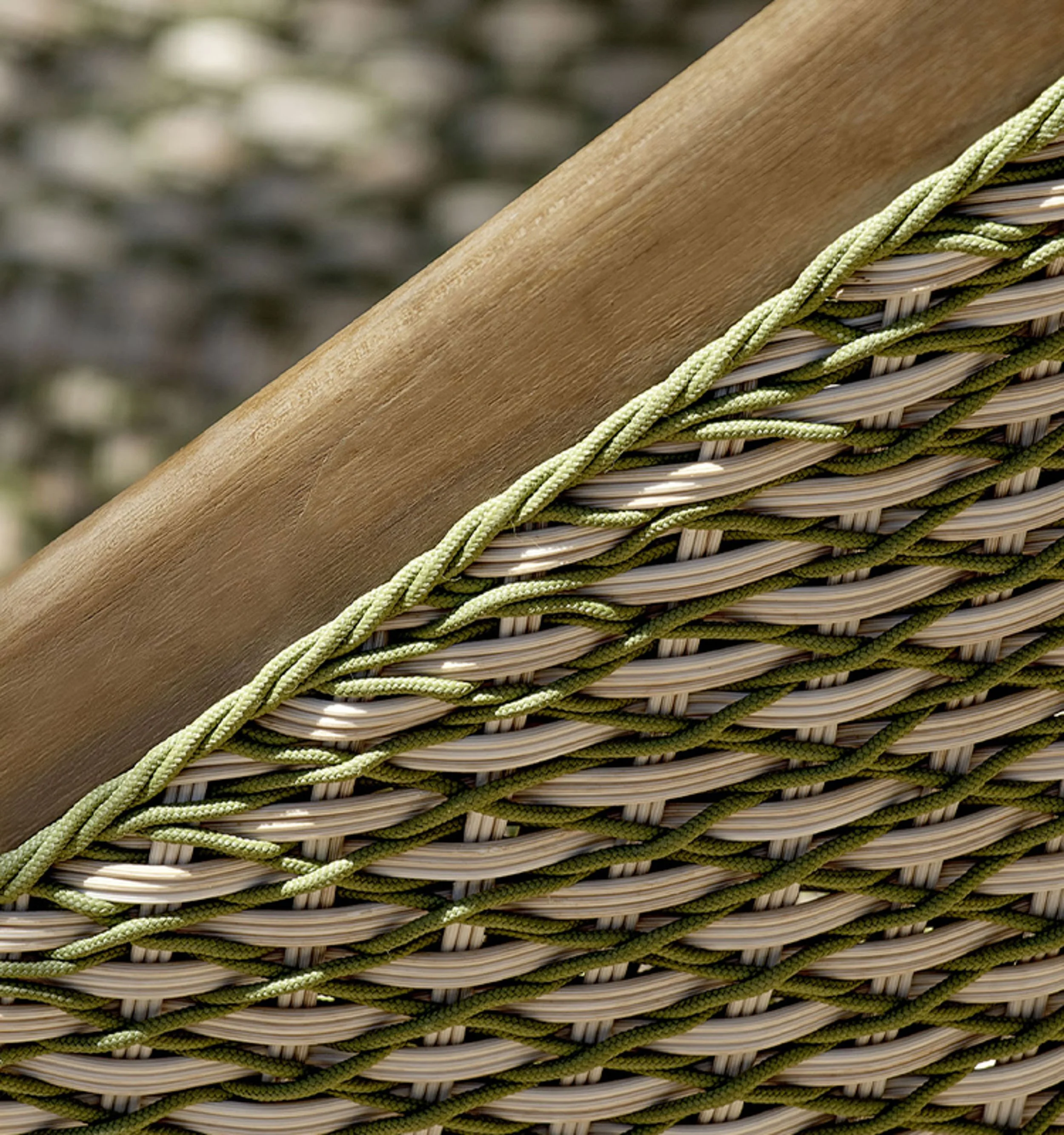 Close-up of woven wicker basket with green and beige ropes intertwined, with a wooden handle and blurred outdoor background.