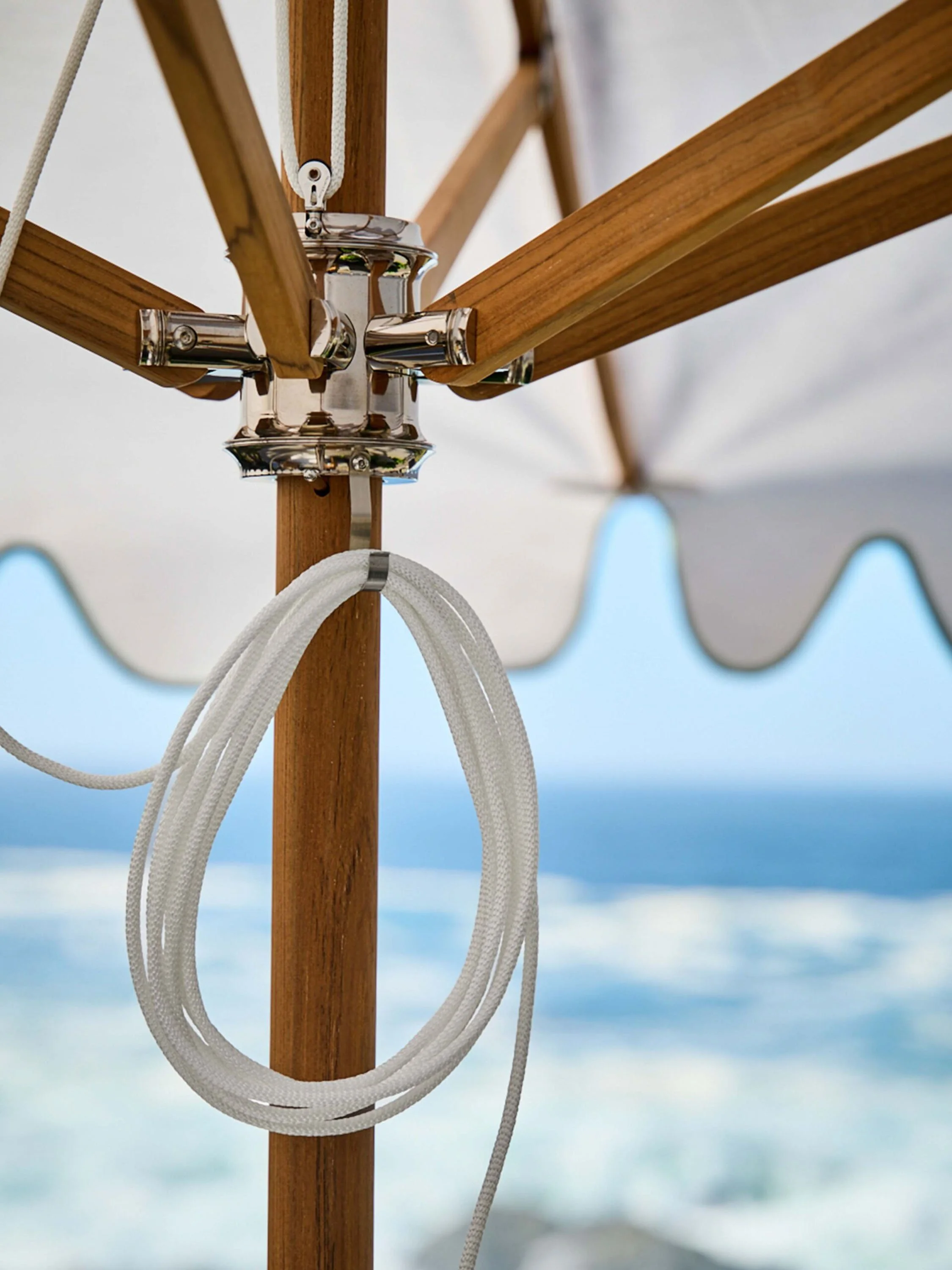 Close-up of a wooden pole with a chrome hardware piece, from which white ropes are hanging, with out-of-focus blue sky and ocean in the background.