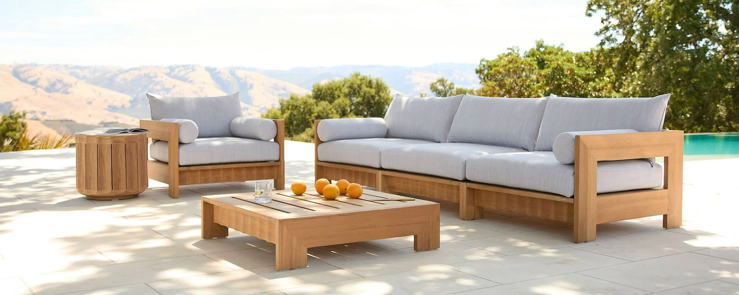 Outdoor patio with wooden furniture, including a sofa, chair, and coffee table, set against a scenic mountain view with trees and a pool to the side.