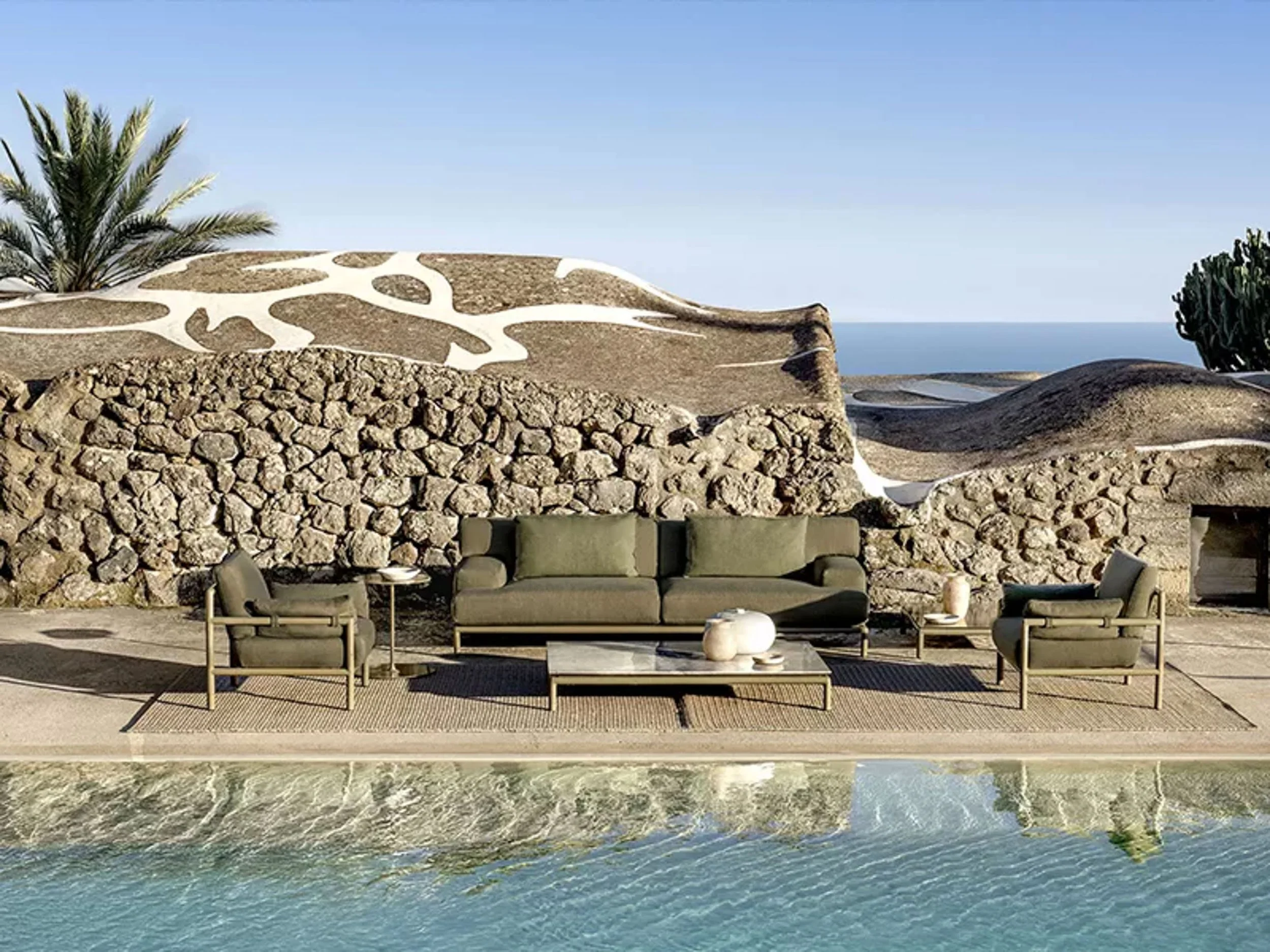 Beachside lounge area with modern seating, stone wall, and scenic view of ocean and cliffs in the background.