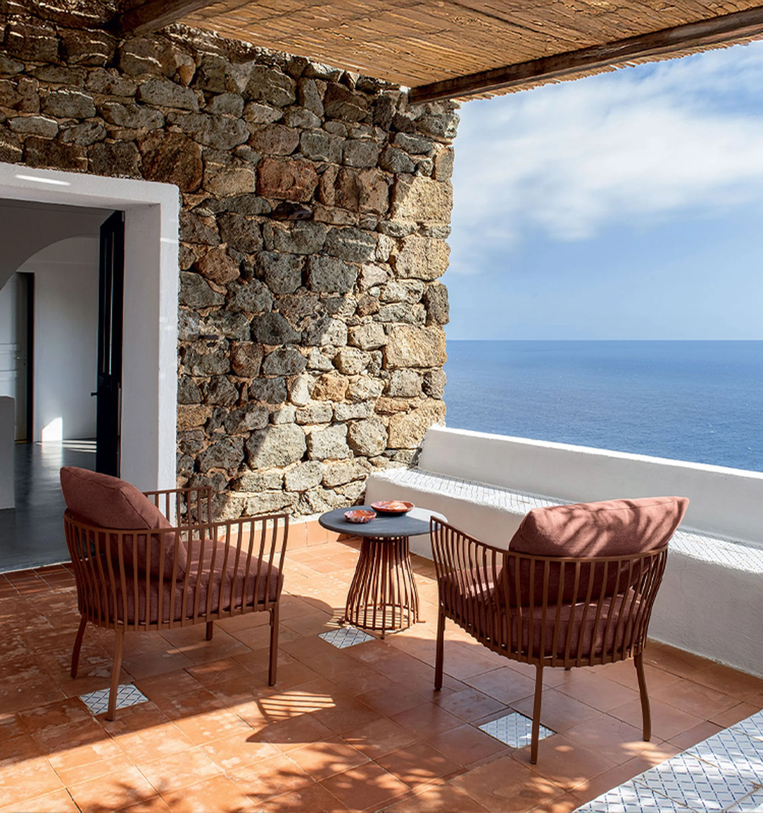 Outdoor patio with terracotta tiles, wooden chairs with pink cushions and a small table, stone wall, open white window, ocean view with blue sky and scattered clouds.