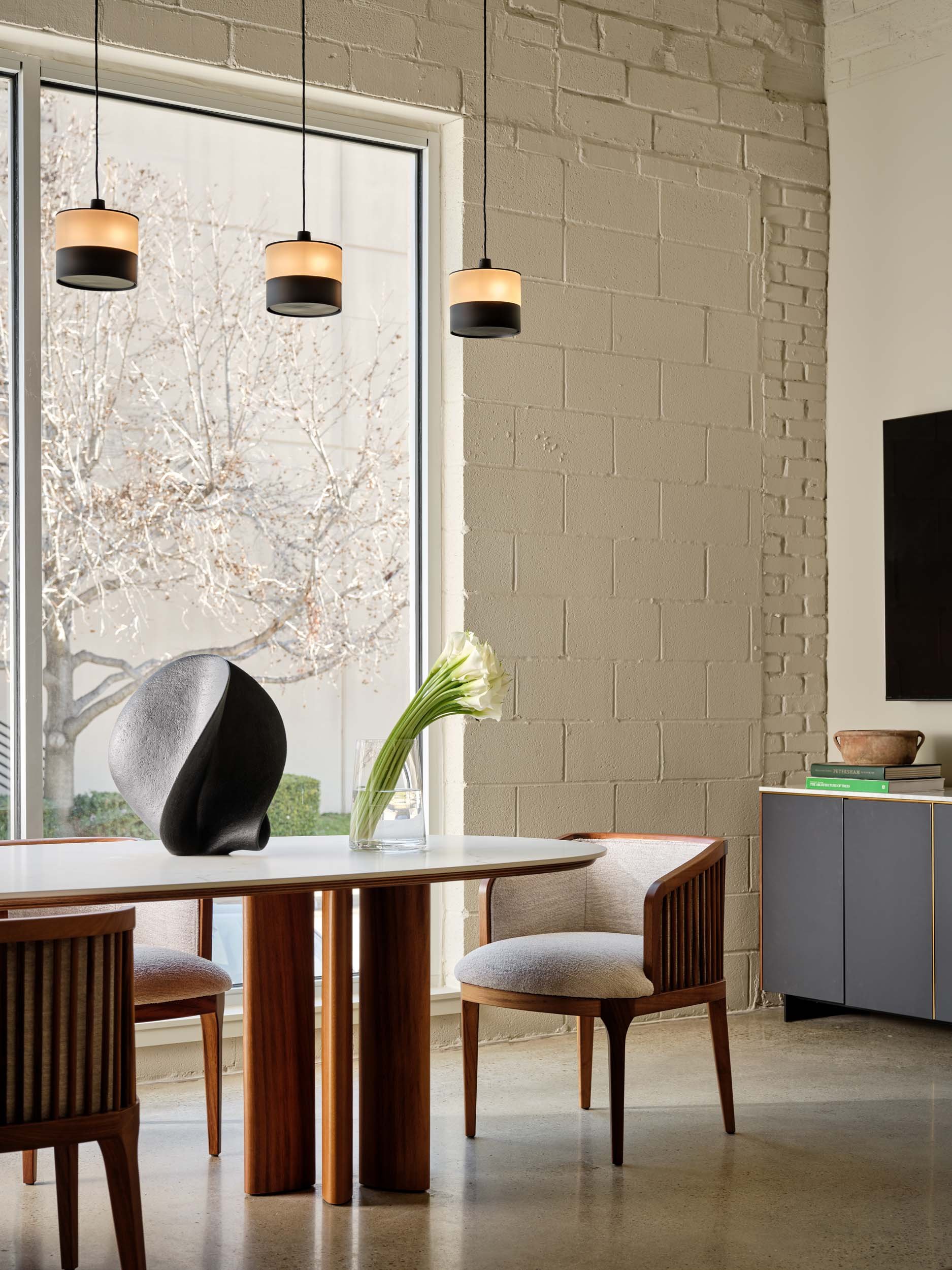 A modern dining room with a wooden table and chairs, a decorative black sculpture, a glass vase with white flowers, large window, and hanging pendant lights.