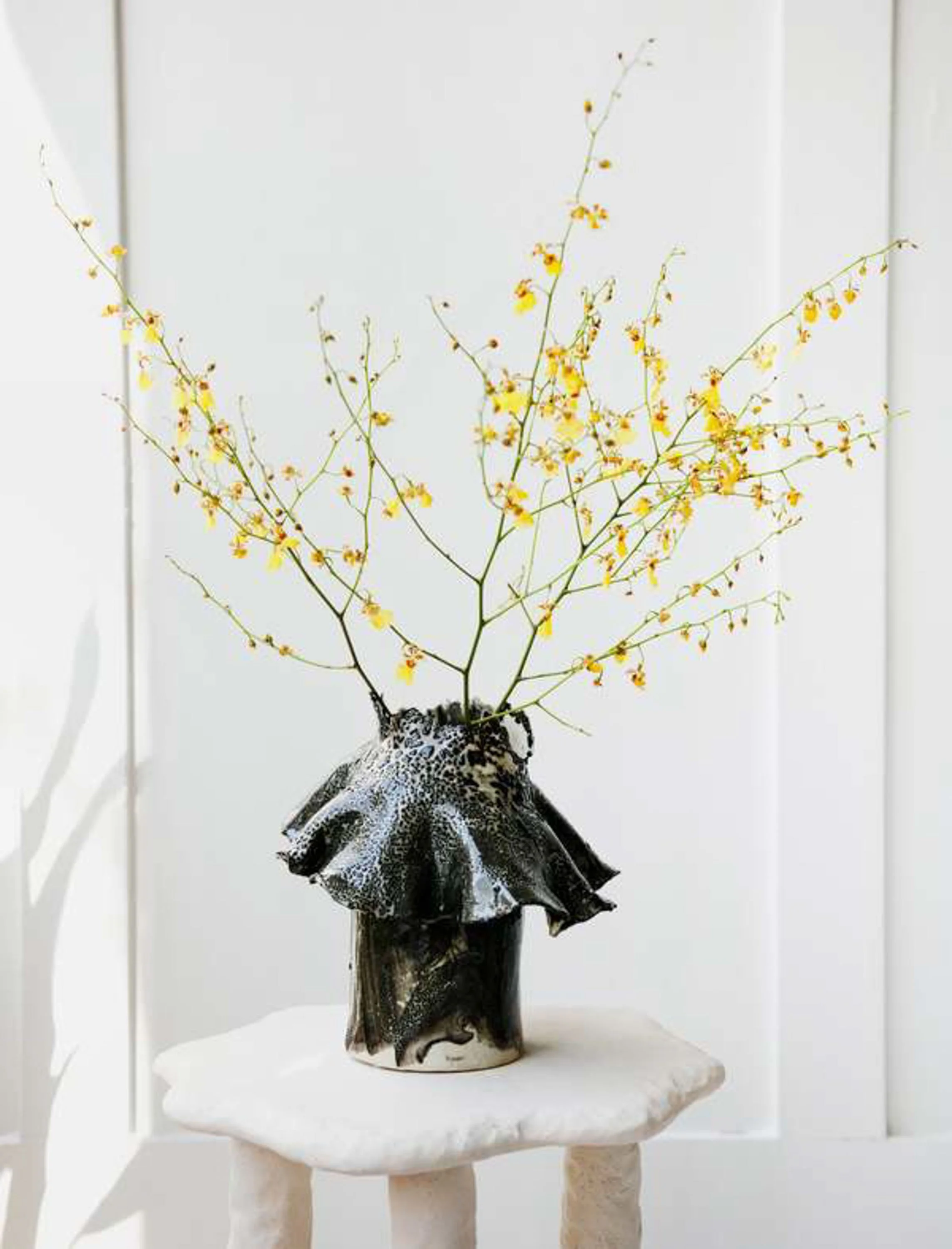 An artistic sculpture of a plant with yellow flowers extending from a black and white vase on a small white table against a plain background.