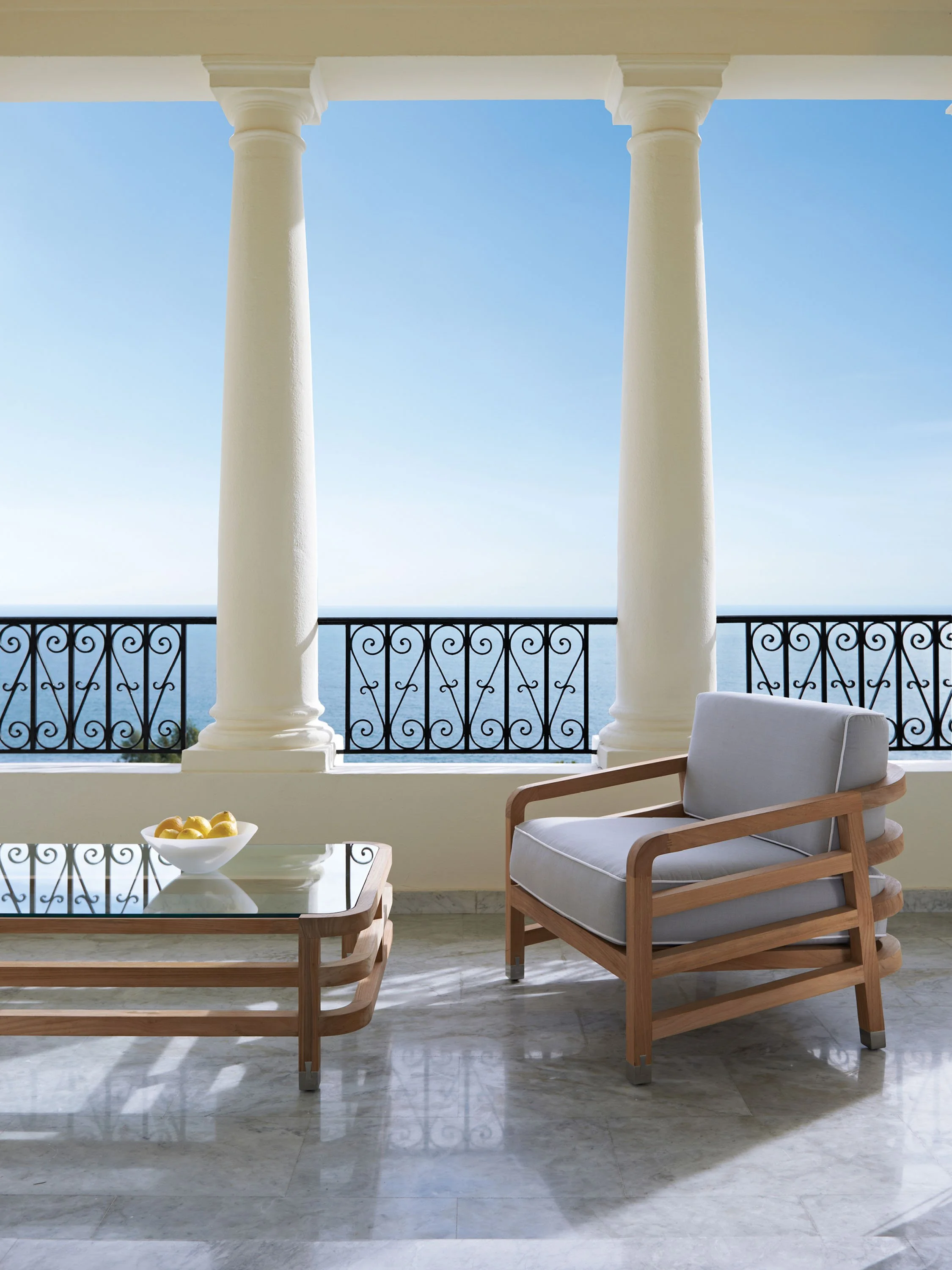 A balcony with two large white columns, a black decorative metal railing, a glass-top wooden table with a bowl of lemons, a modern wooden armchair with light gray cushions, and a view of the ocean under a clear blue sky.