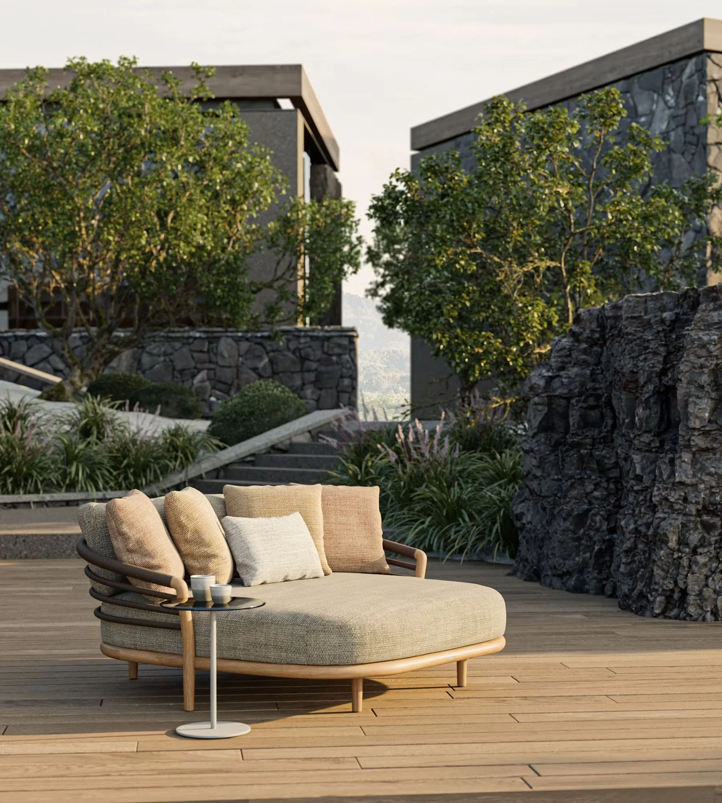 Modern outdoor patio with a curved beige cushioned sofa, pillows, a small round side table with two cups, and greenery including trees and shrubs, on a wooden deck with a stone wall and contemporary buildings in the background.