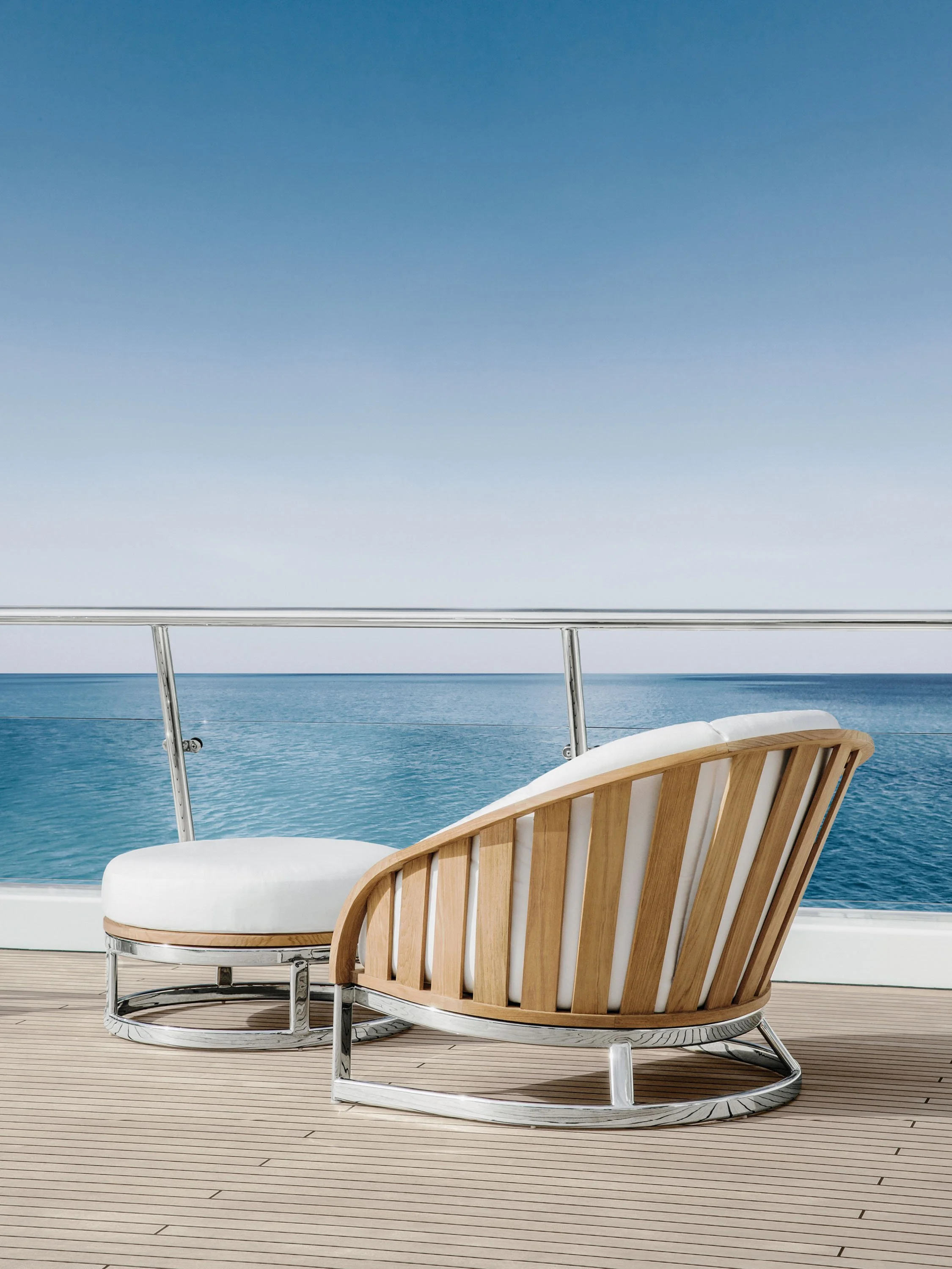 Two modern wooden chairs with white cushions on a deck overlooking the ocean under a clear blue sky.