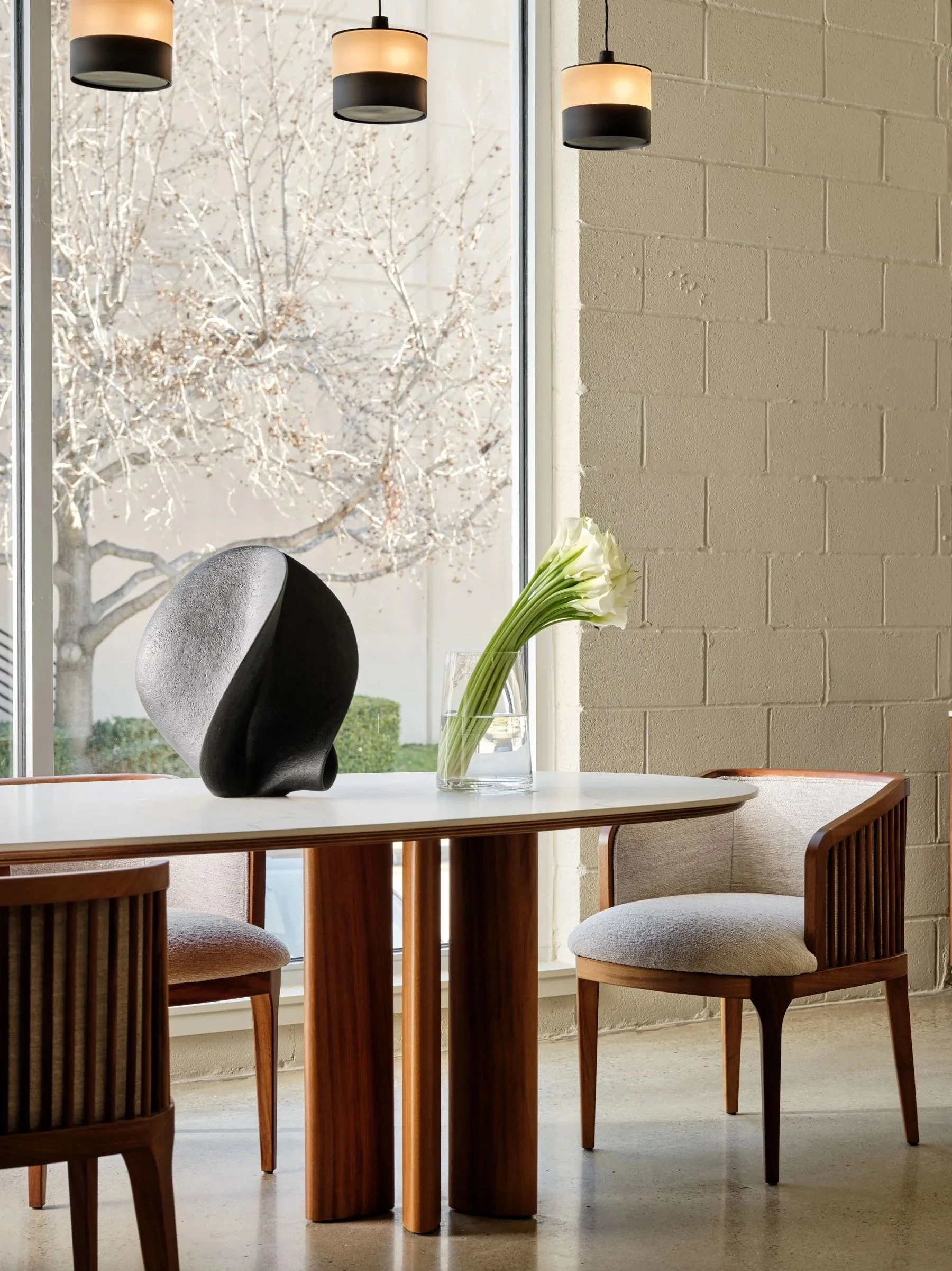 Interior of a modern dining area with a white table, wooden chairs, a glass vase with white calla lilies, and black pendant lights. Large window showing leafless trees outside.