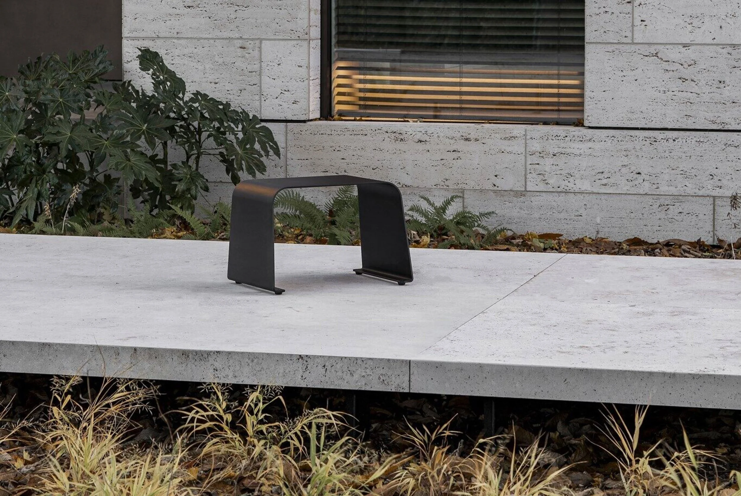 Modern building exterior with a window with horizontal blinds, a black bench on a concrete surface, and plants with green and brown leaves in the foreground.