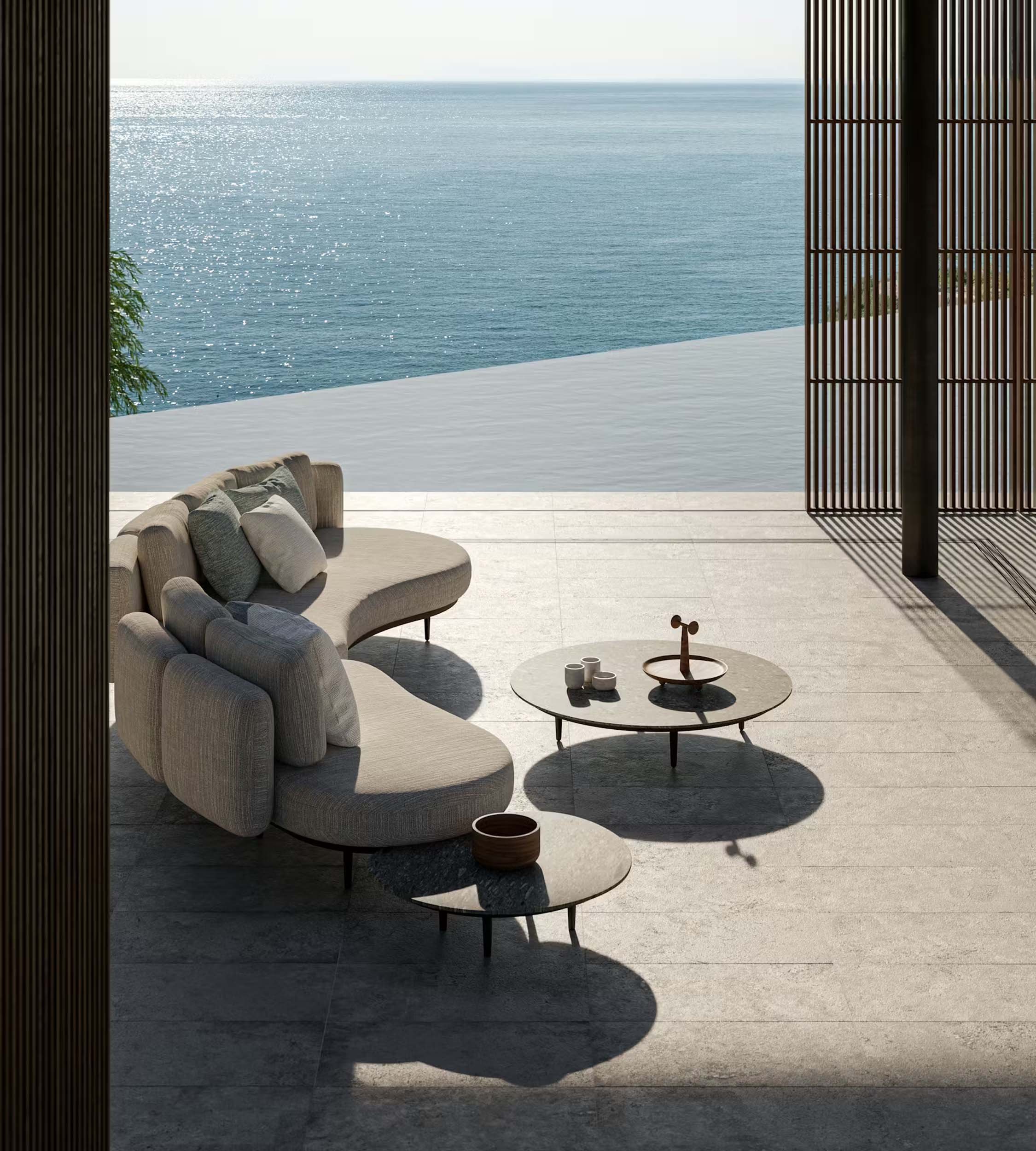 View from a shaded outdoor patio overlooking the ocean with a sitting area, including a cushioned sofa, a small round table with decorative items, and wood slatted partitions casting shadows.
