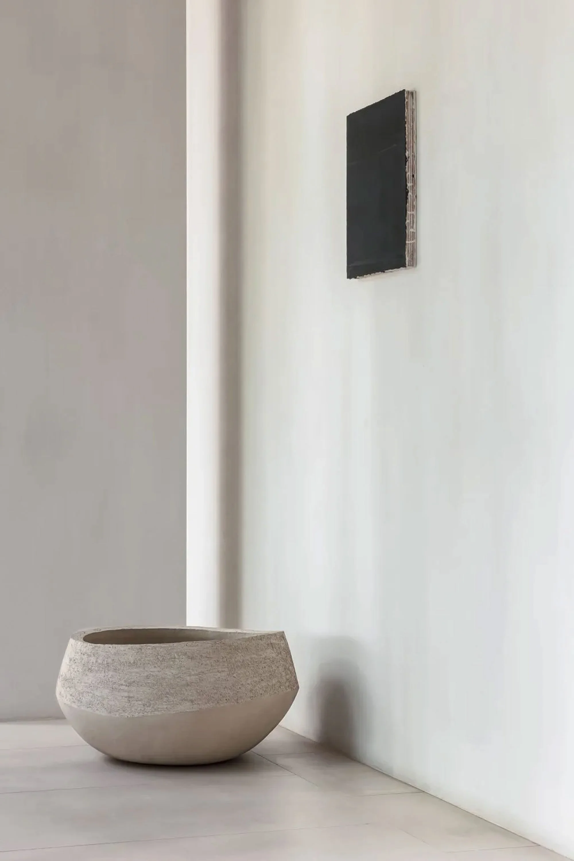 Large, round, textured, light-colored ceramic planter on the floor, with a tall, white wall behind it. A small, black rectangular object or artwork is mounted on the wall.