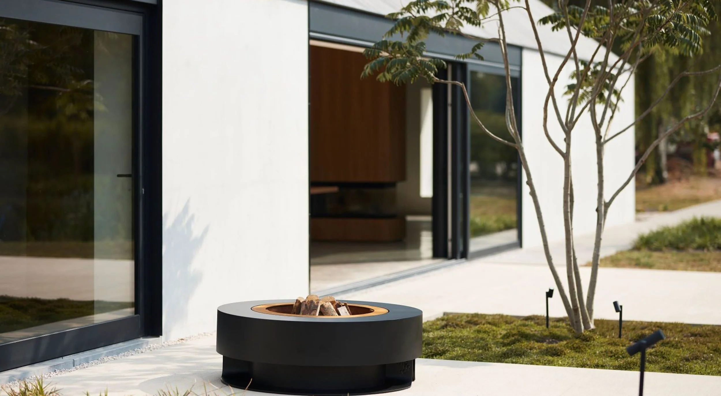 Modern house with large glass sliding doors, small tree, and a black fire pit with wood inside outside on a patio.