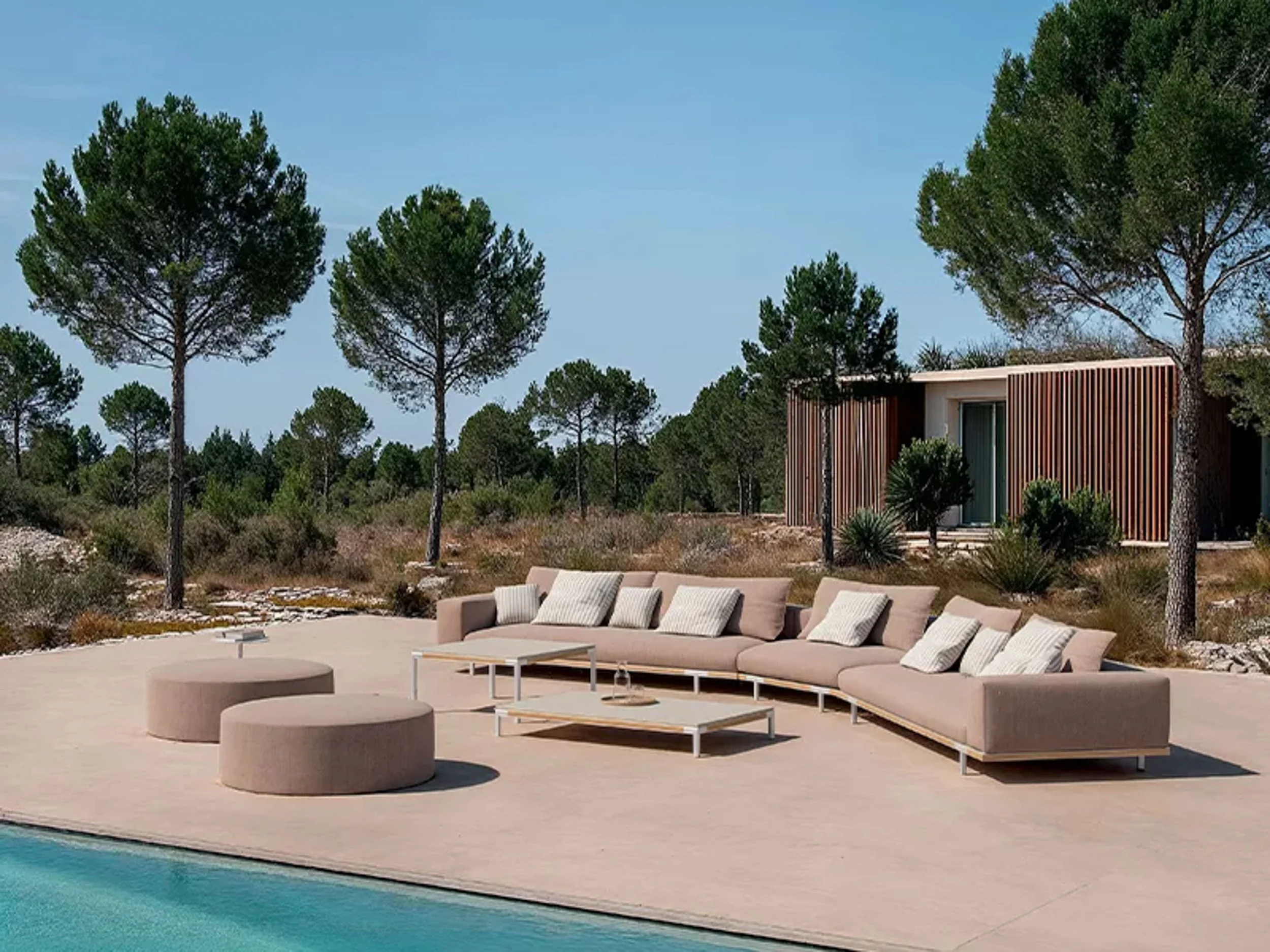 Modern outdoor seating area with beige sectional sofa, white tables, and cylindrical ottomans near a swimming pool, surrounded by pine trees and a contemporary building with vertical wood slats.