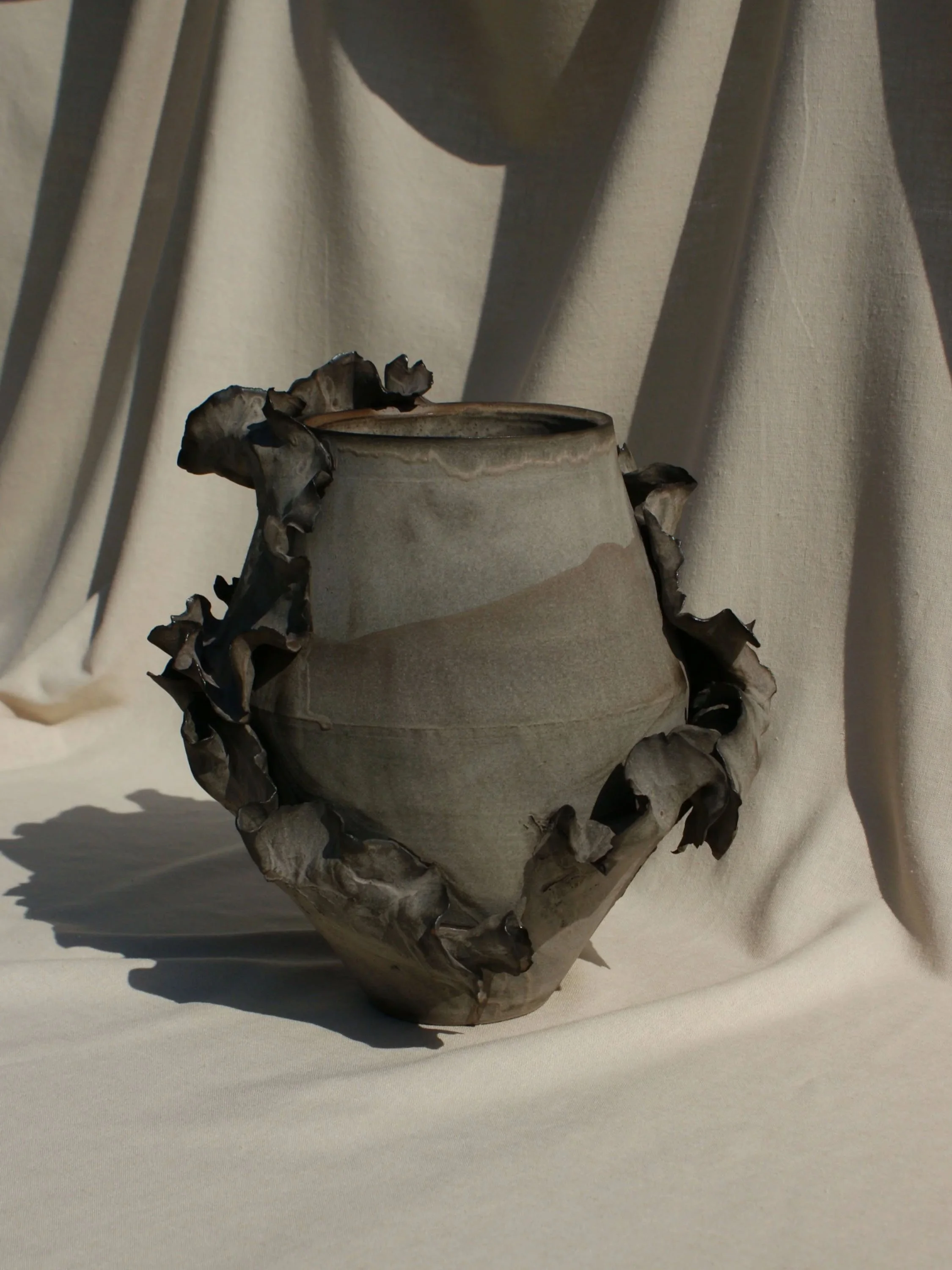A ceramic vessel with a wavy, ruffled rim and leaf-like handles, placed on a neutral fabric background.
