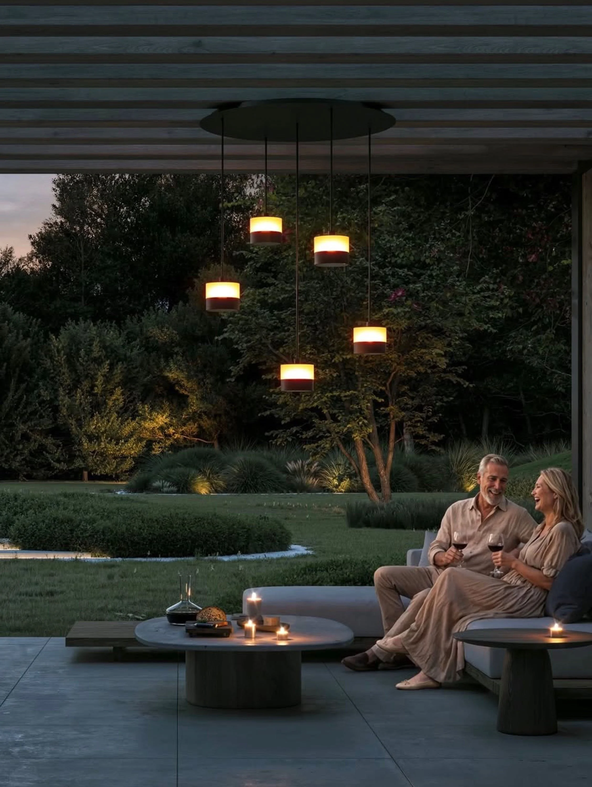 A couple sitting on an outdoor sofa, holding glasses of red wine, and smiling at each other during the evening. There are candles on the table in front of them, and a background of trees and sky with outdoor hanging lights.