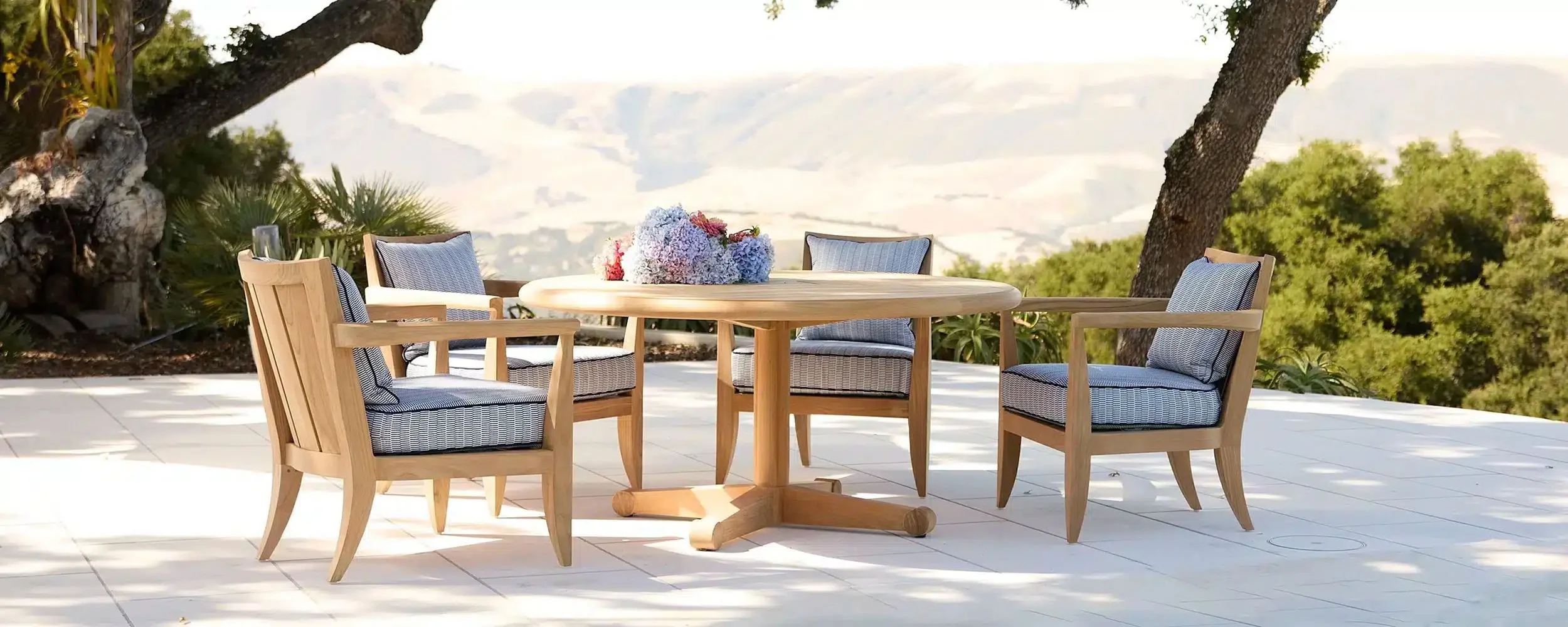 Outdoor patio with a wooden round table and six chairs with gray cushions, set on white tiled flooring, with trees and green foliage in the background, and a mountain range visible in the distance.