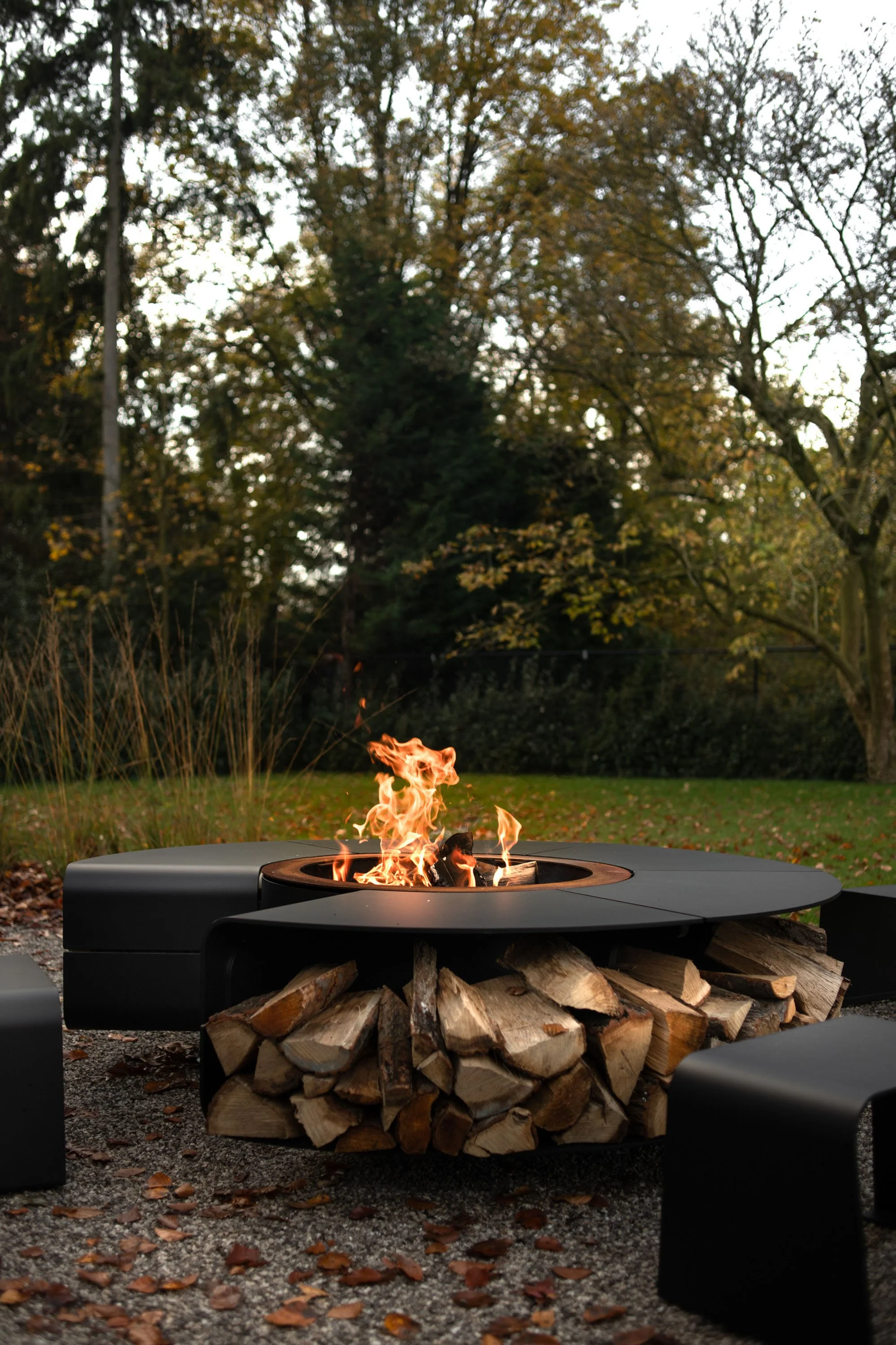 Fire pit with wood logs underneath and a small flame burning, set outdoors with a background of trees and a cloudy sky.