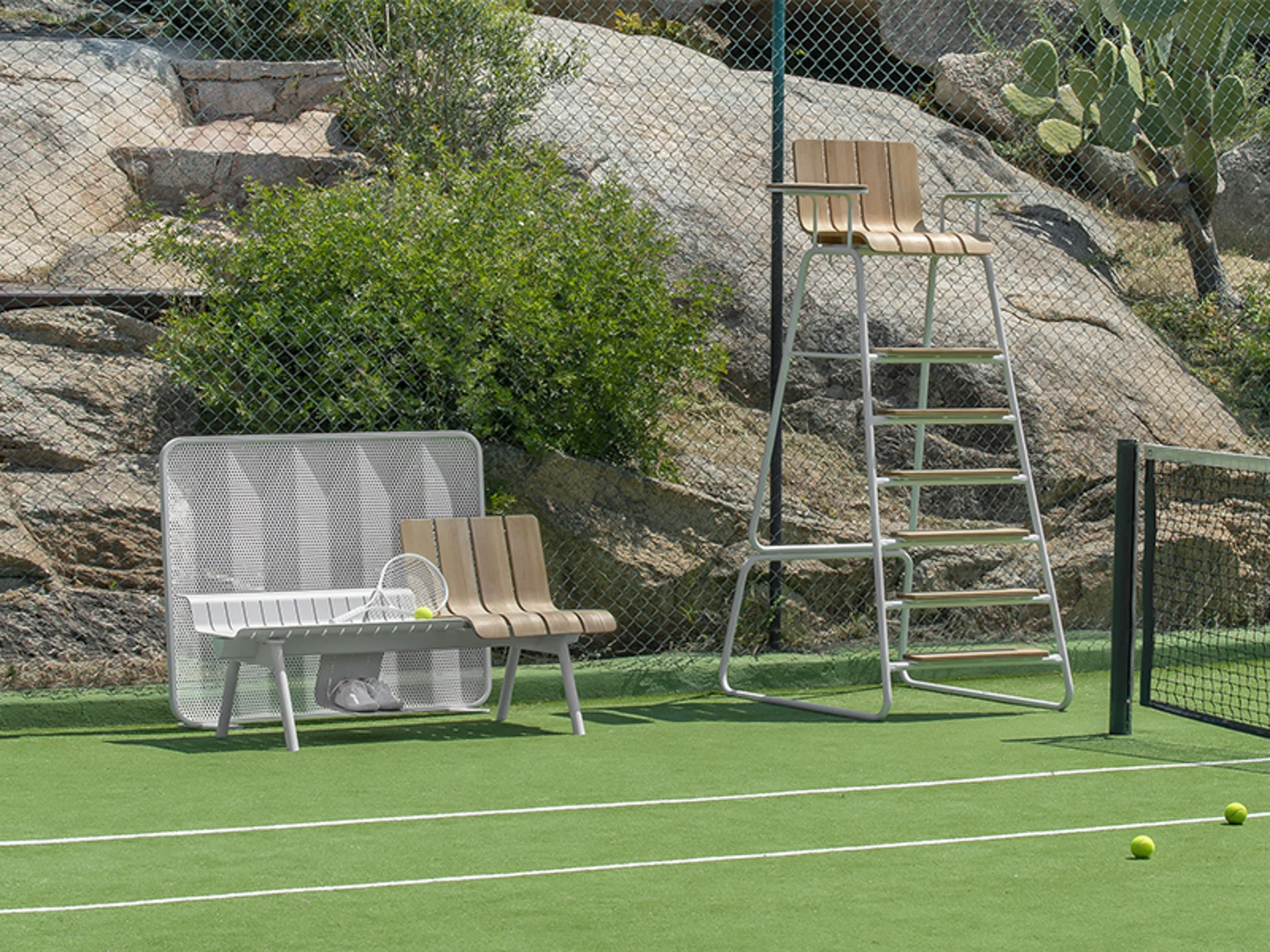 A tennis court with green surface, a white bench with tennis shoes, two tennis balls, a tan chair, a beige tall ladder, a metal fence, rocks, dirt, and cactus plants in the background.