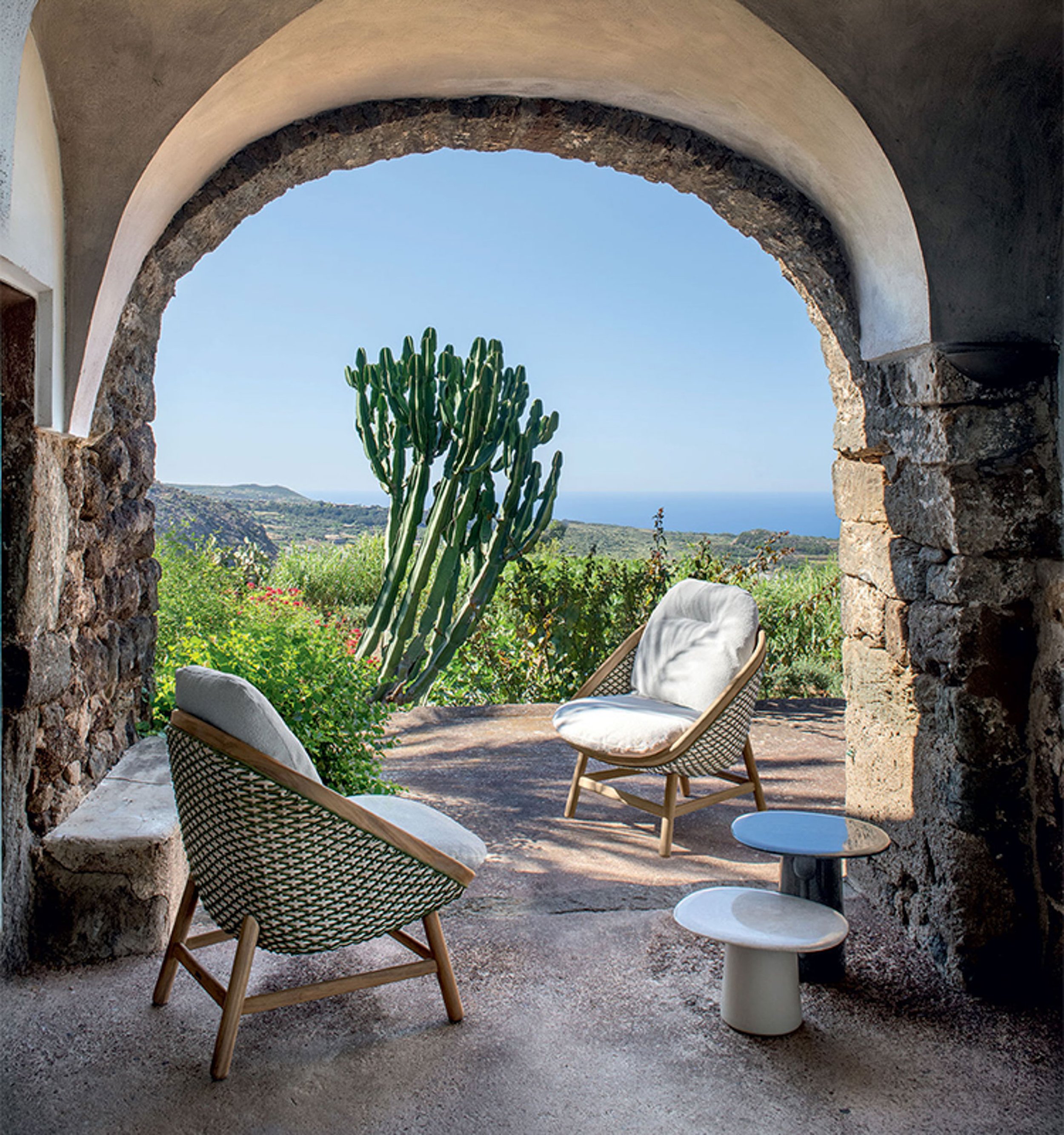 View through a stone archway to a patio with two patterned chairs and a small round side table. Beyond the arch, there are green shrubs, a cactus, and a distant landscape with hills and the ocean.