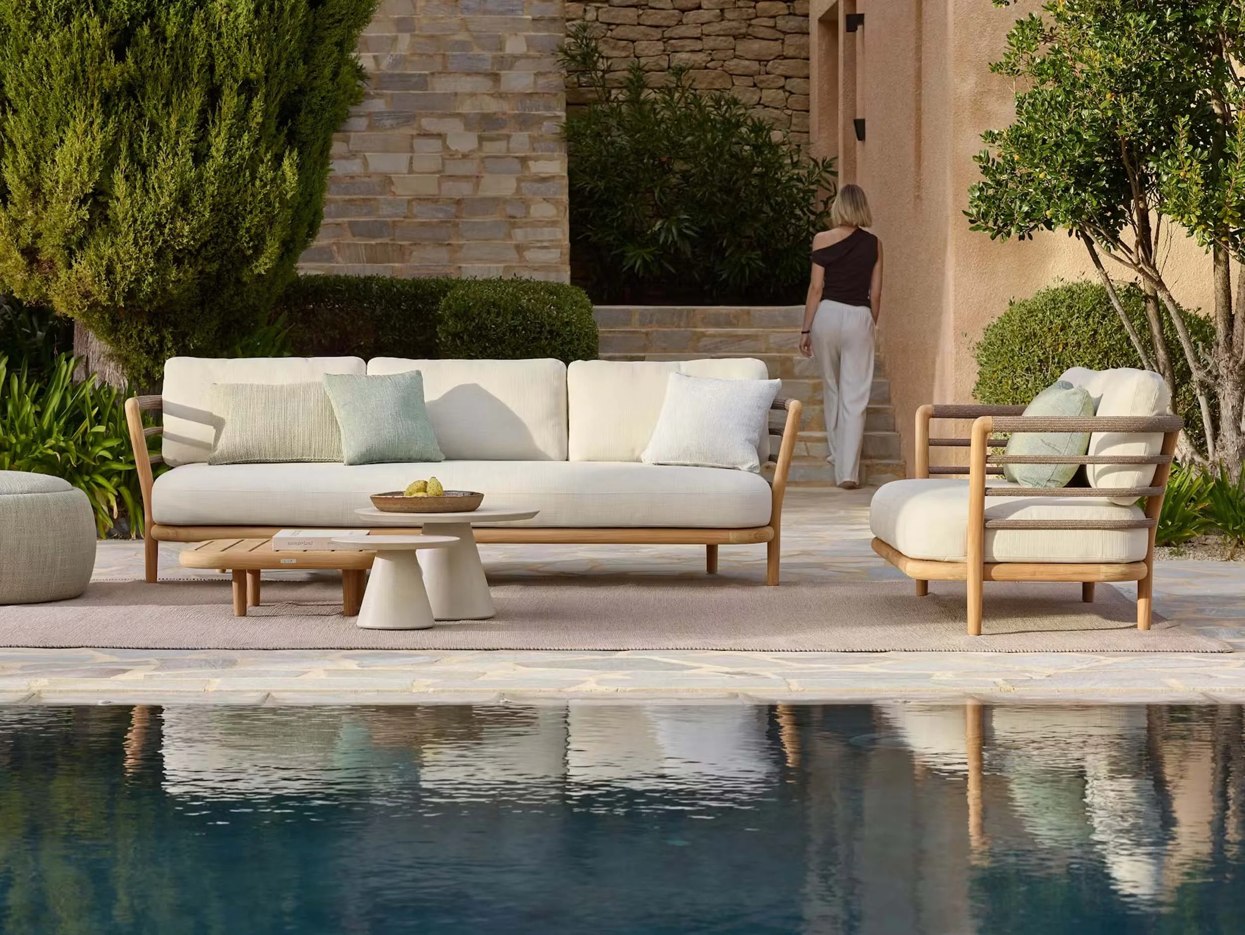 An outdoor patio area with a swimming pool in the foreground, a white sofa with pillows, and a woman walking up stone stairs towards a house. The scene is surrounded by lush green trees and bushes, with a stucco wall and stone accents.