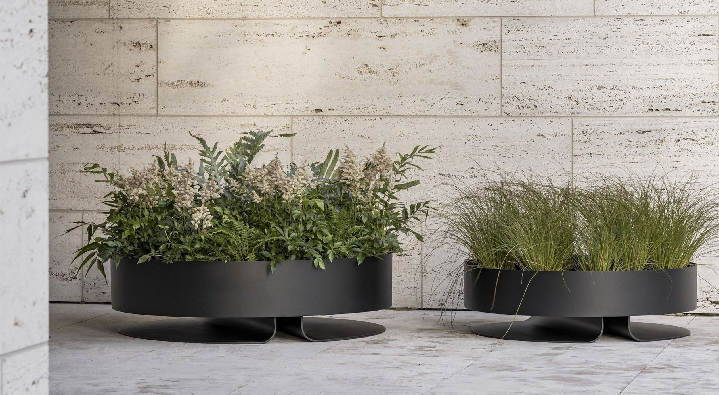 Two black rectangular planters with various green and white plants, placed against a white textured brick wall.
