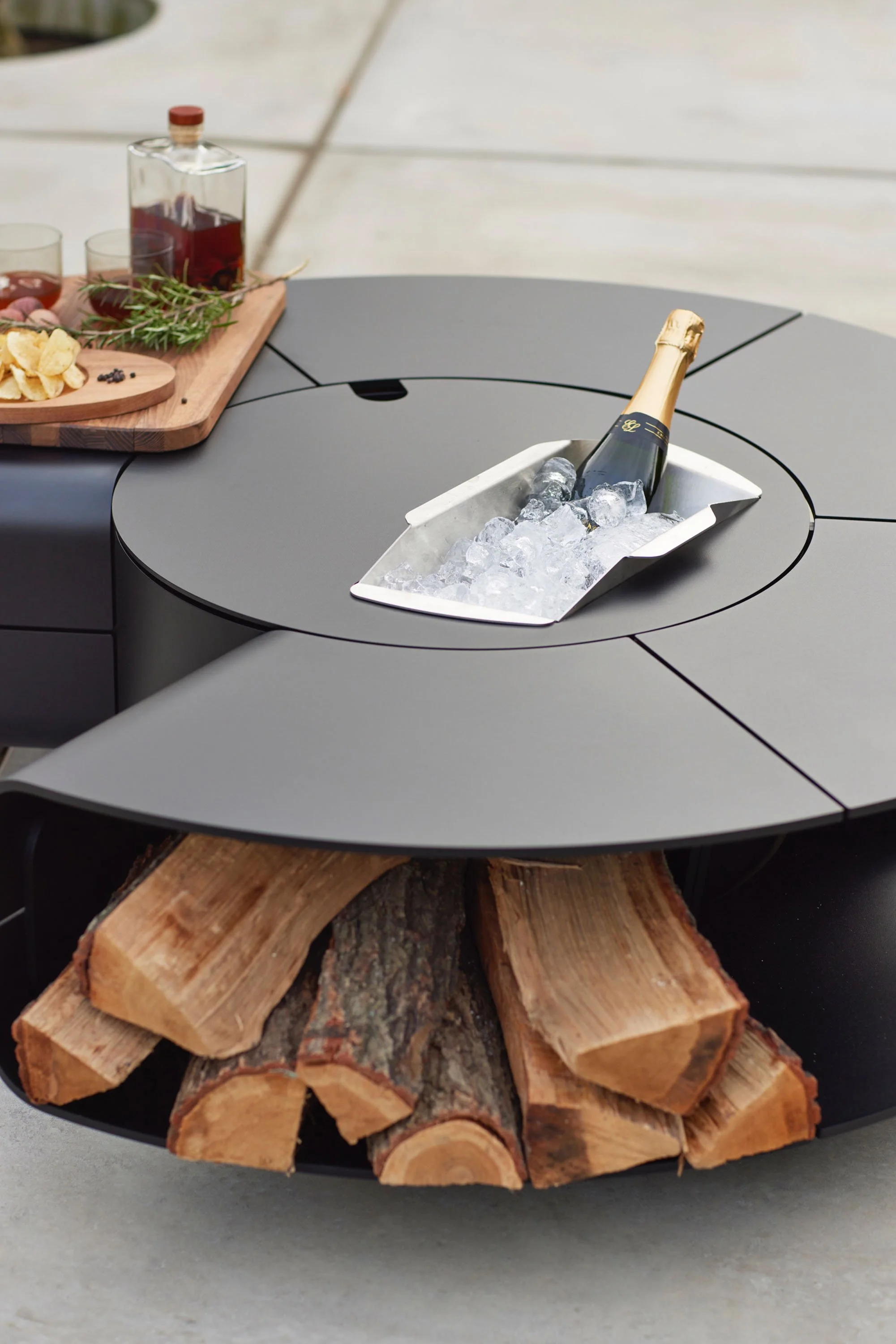 A bottle of champagne in a metal ice bucket filled with ice, placed on a black table with a rustic wooden log underneath. In the background, there is a wooden tray with food and drink items and some green herbs.