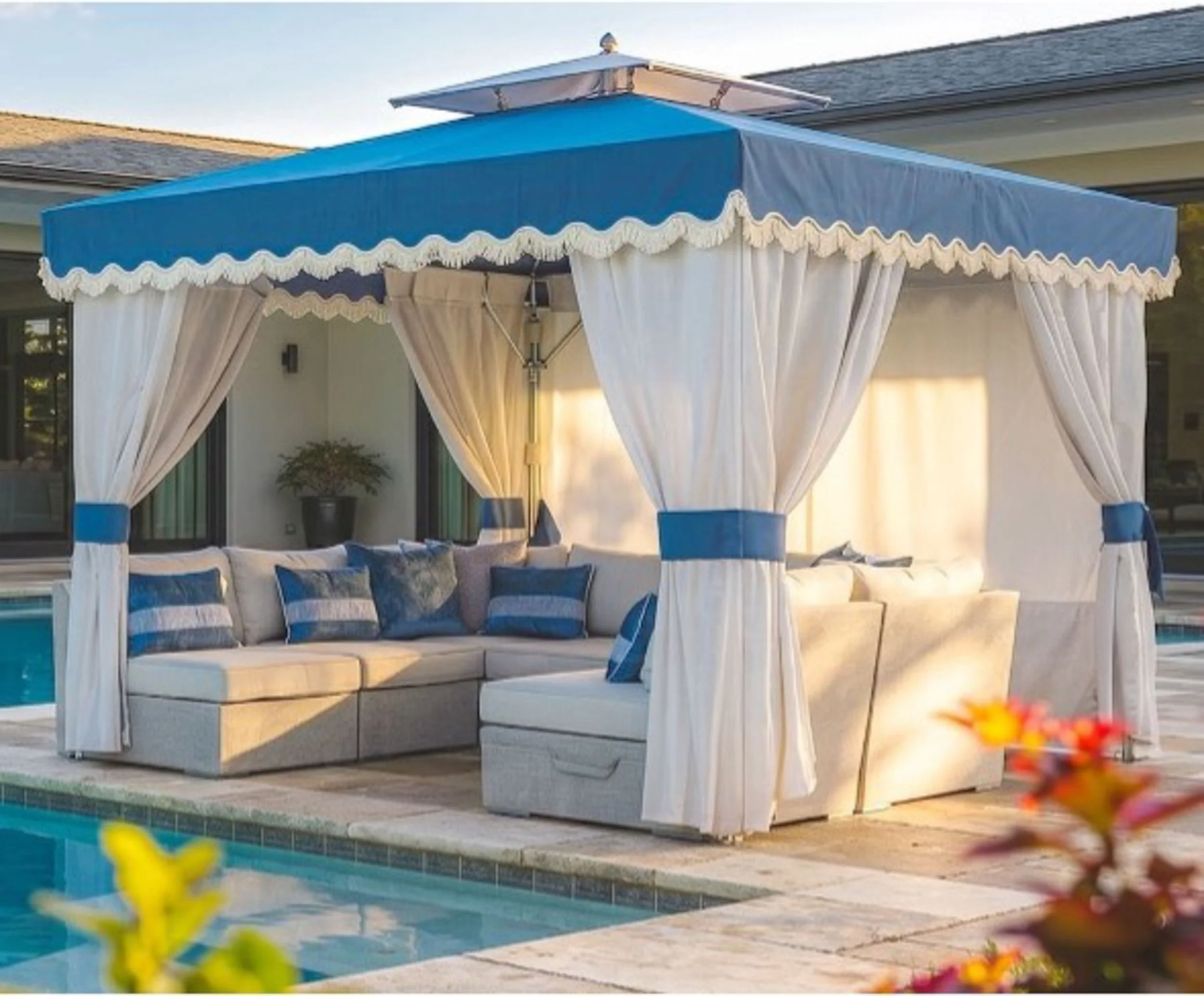 A blue and white cabana with curtains, set up next to a swimming pool at a house with a gray roof in the background.