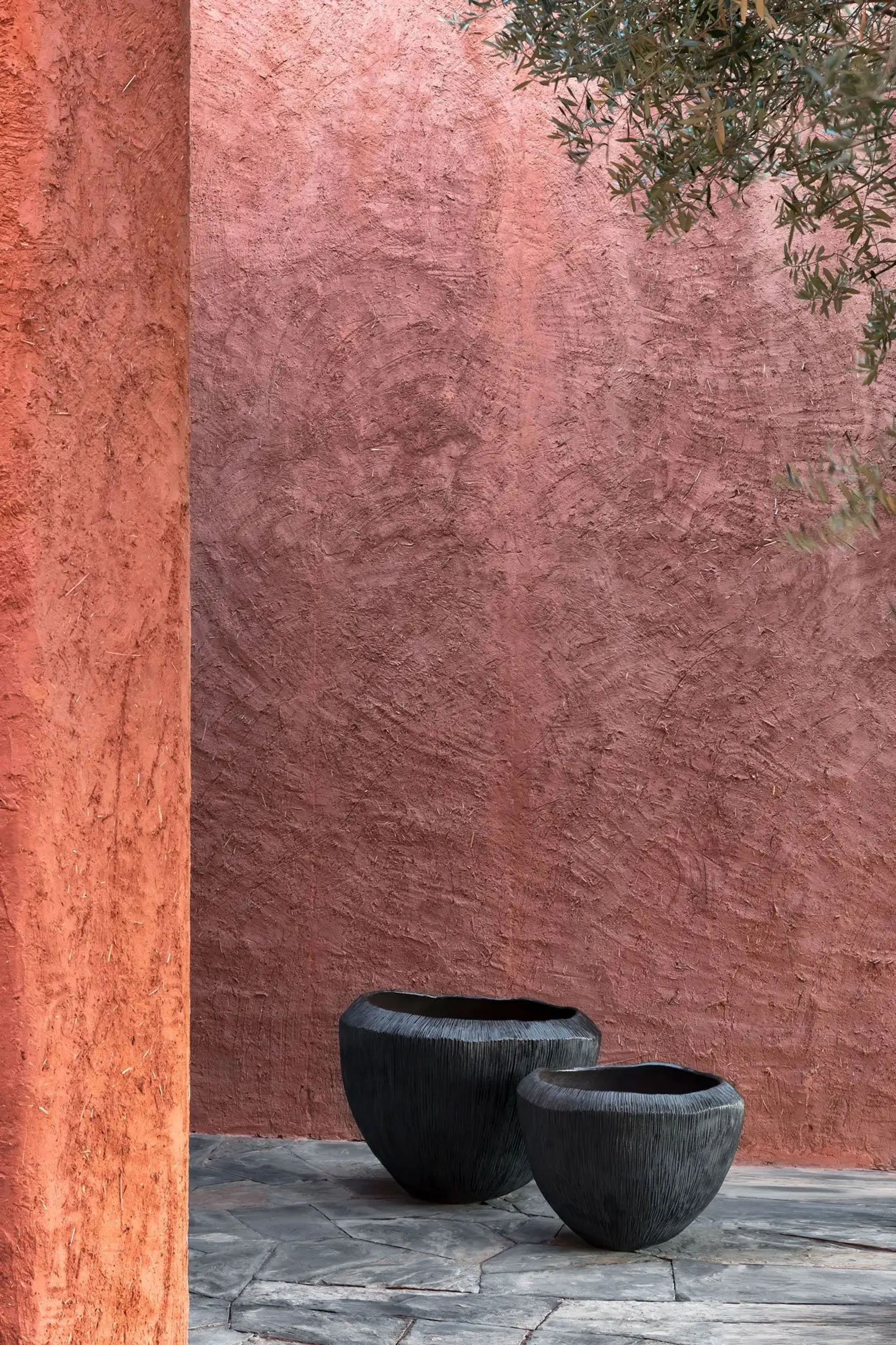 Two black textured ceramic or stone pots on a grey stone pavement, placed against a pinkish-red stucco wall, with green foliage partly visible on the upper right.