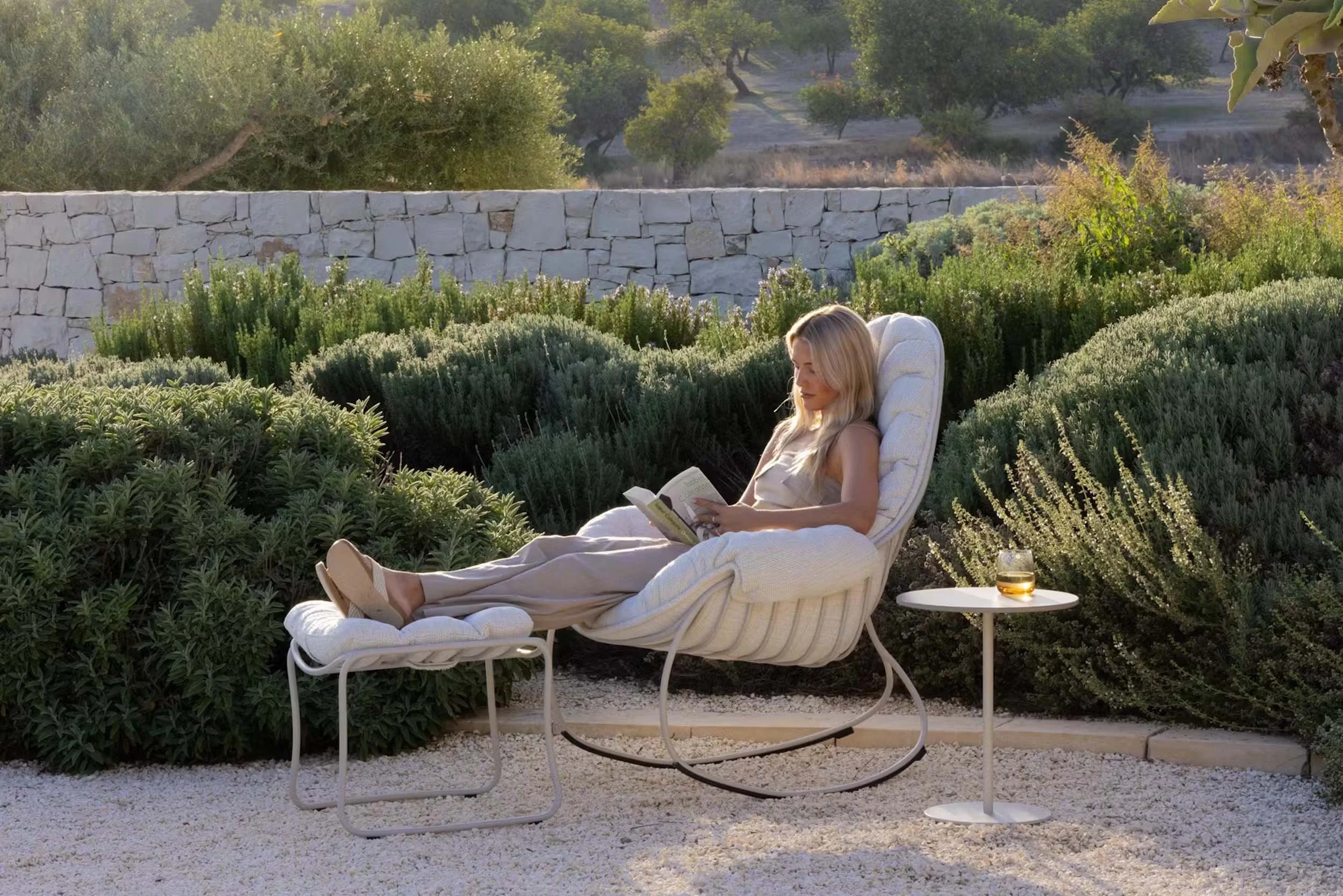 A woman relaxing outdoors on a lounge chair, reading a book, with a glass of whiskey and ice nearby, surrounded by lush green bushes and trees in the background.