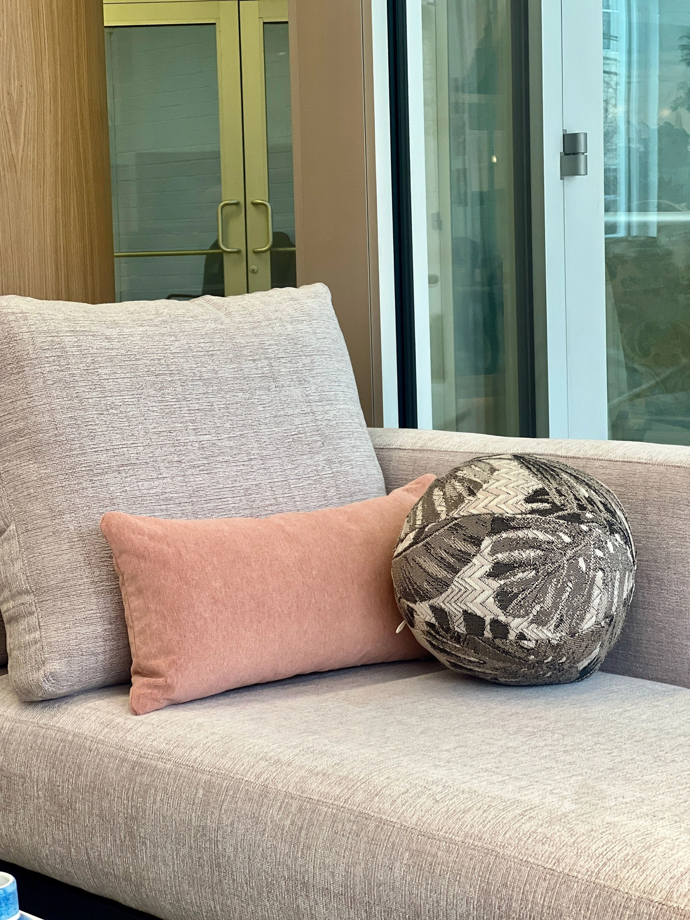 Close-up of a beige fabric sofa with a pink rectangular pillow and a round decorative pillow with a black, cream, and gray abstract pattern. Behind the sofa, there is a large window with a sliding glass door, and a reflection of a plant is visible outside.