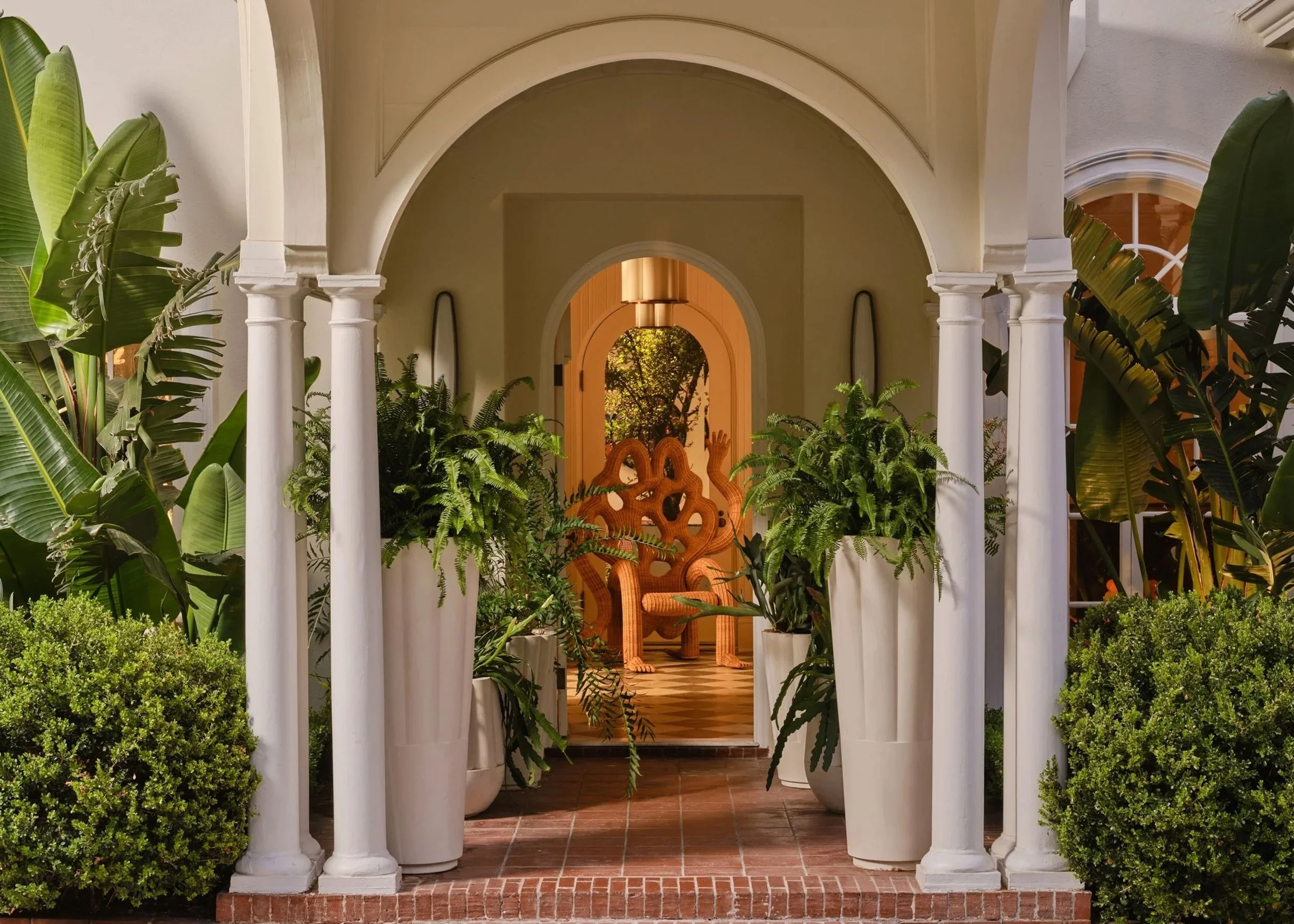 An elegant doorway framed by white columns with lush greenery on either side, leading to a cozy, warmly lit interior with wooden accents and decorative wooden chairs.