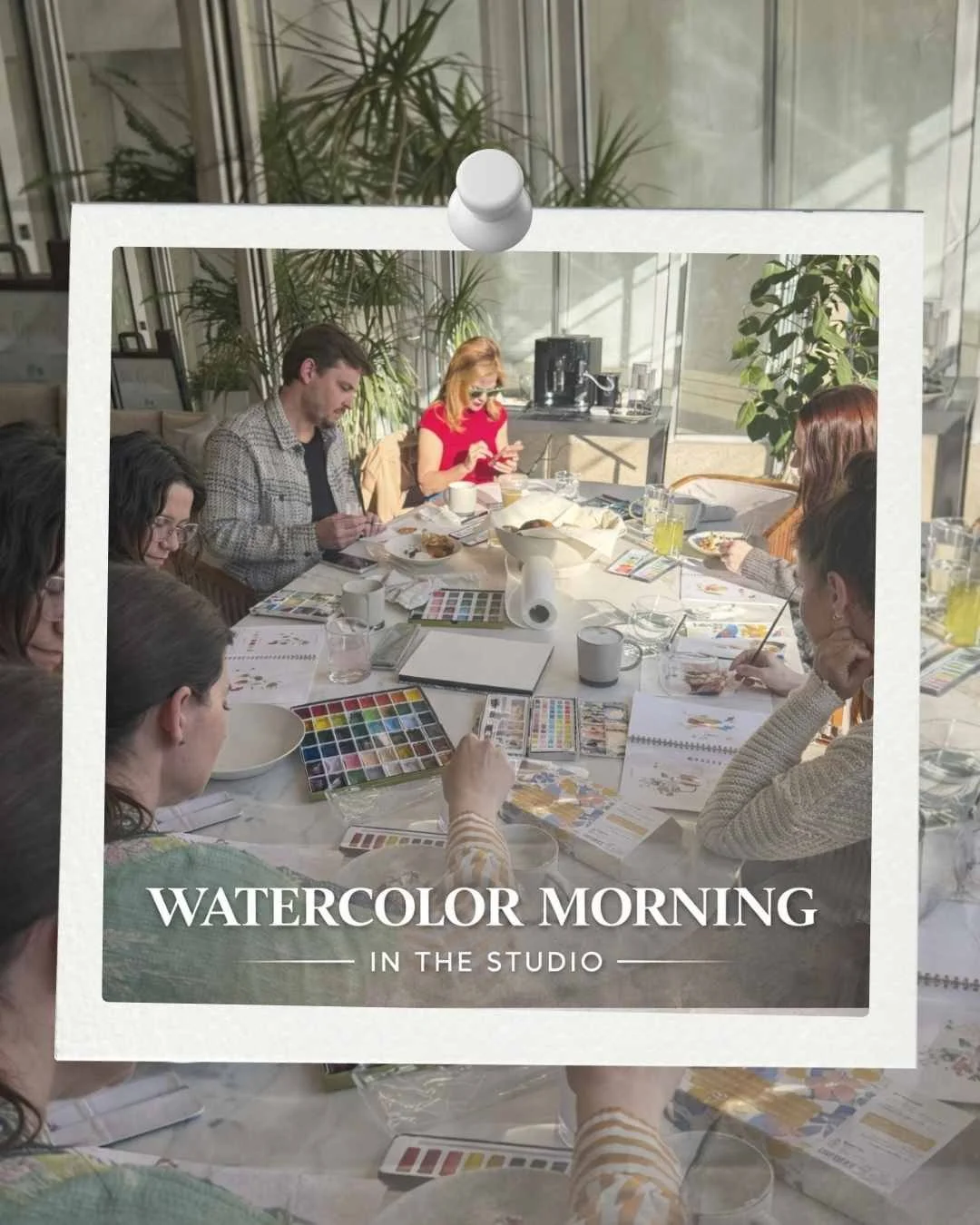 A group of people sitting around a table with watercolor painting supplies, engaging in a watercolor morning class in a bright studio with large windows and plants.